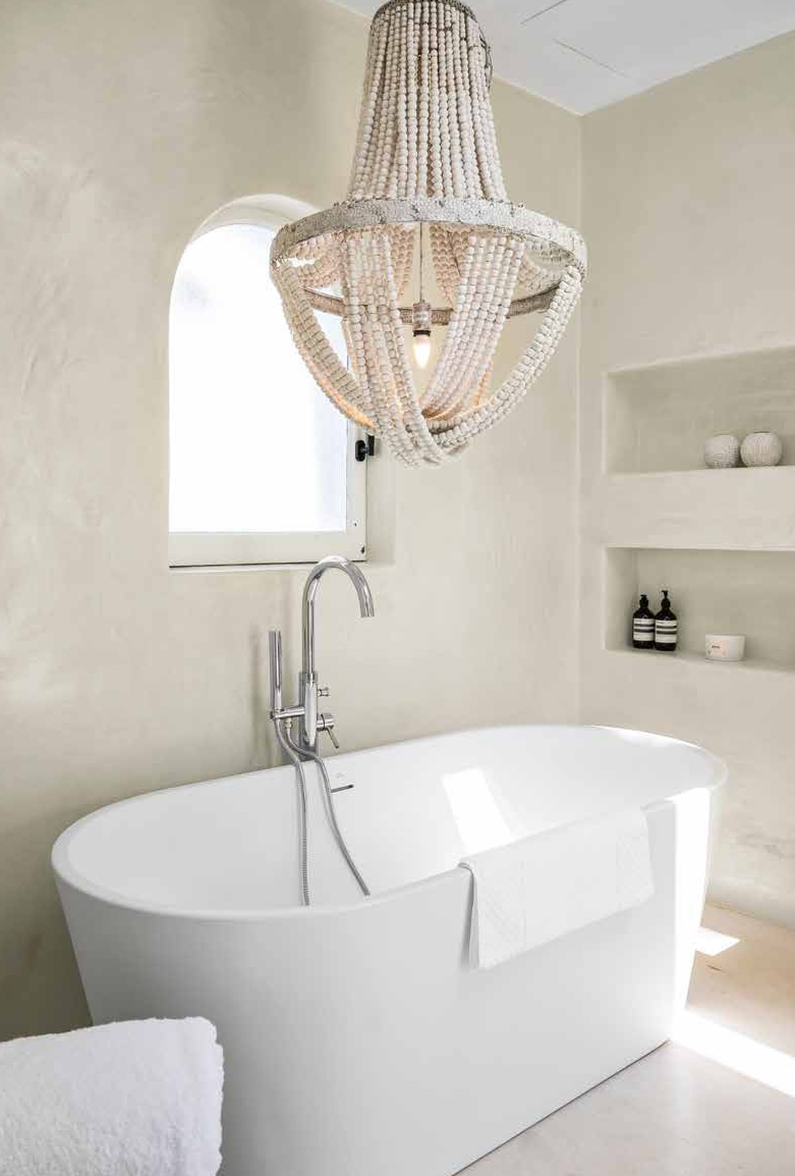 White freestanding bathtub in a minimalist bathroom with a beaded chandelier hanging above, a small window, and built-in shelves with toiletries.