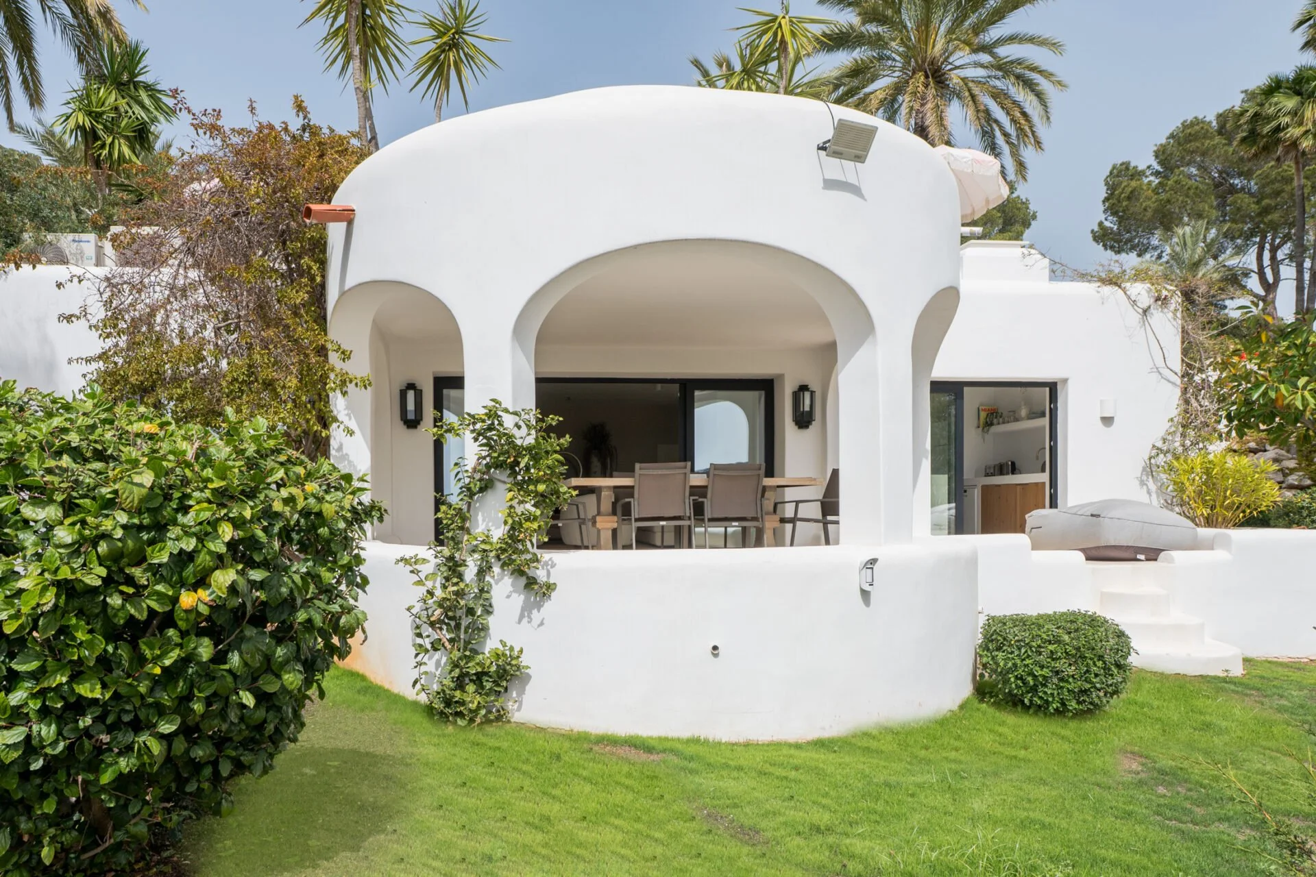 White modern house with rounded archways, outdoor patio with dining table and chairs, surrounded by greenery and palm trees.