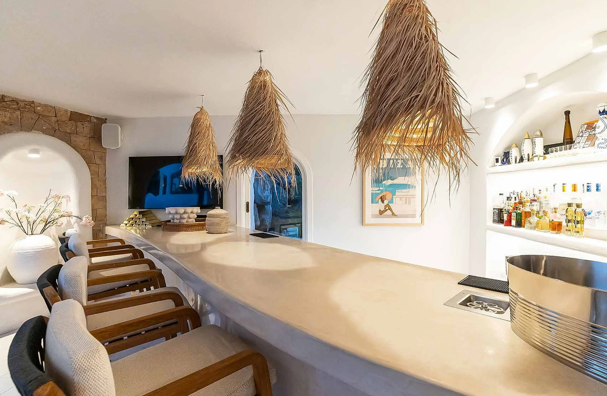 Bar counter with wooden chairs, hanging straw pendant lights, a framed beach-themed picture on the wall, and liquor bottles on illuminated shelves.