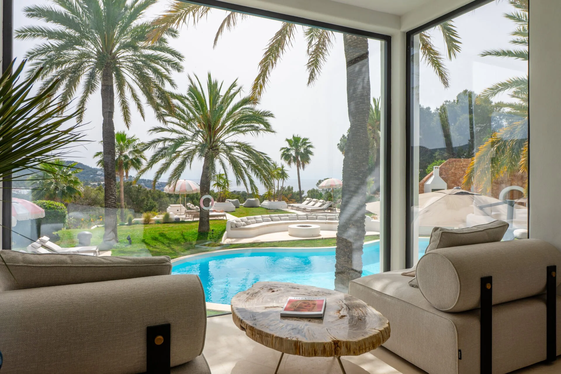 Living room with large glass windows looking out onto a backyard with palm trees, lounge chairs, umbrellas, a lawn, and a swimming pool.