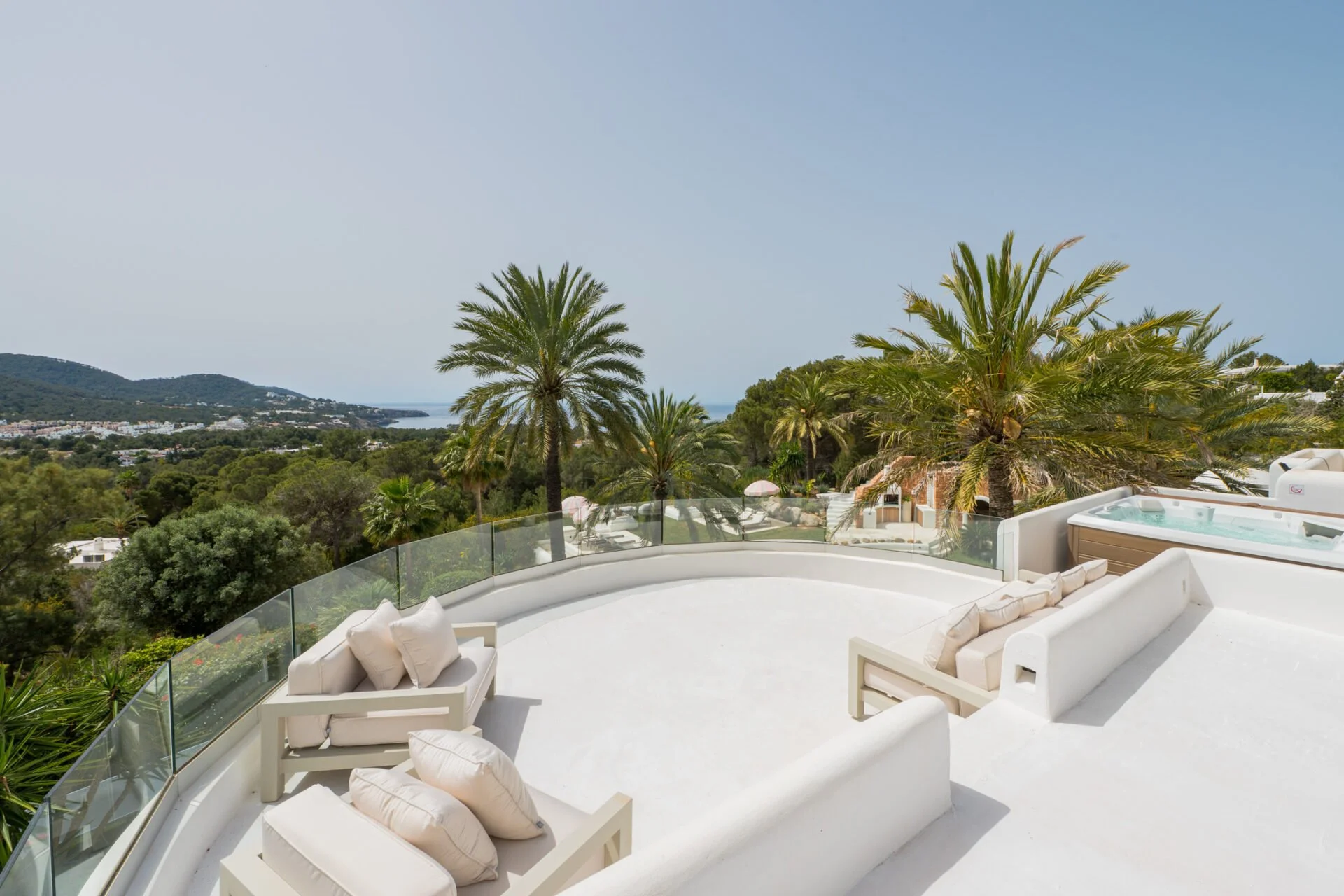 Luxury outdoor terrace with white furniture and a hot tub, overlooking a lush landscape with palm trees, hills, and the sea in the distance under a clear blue sky.
