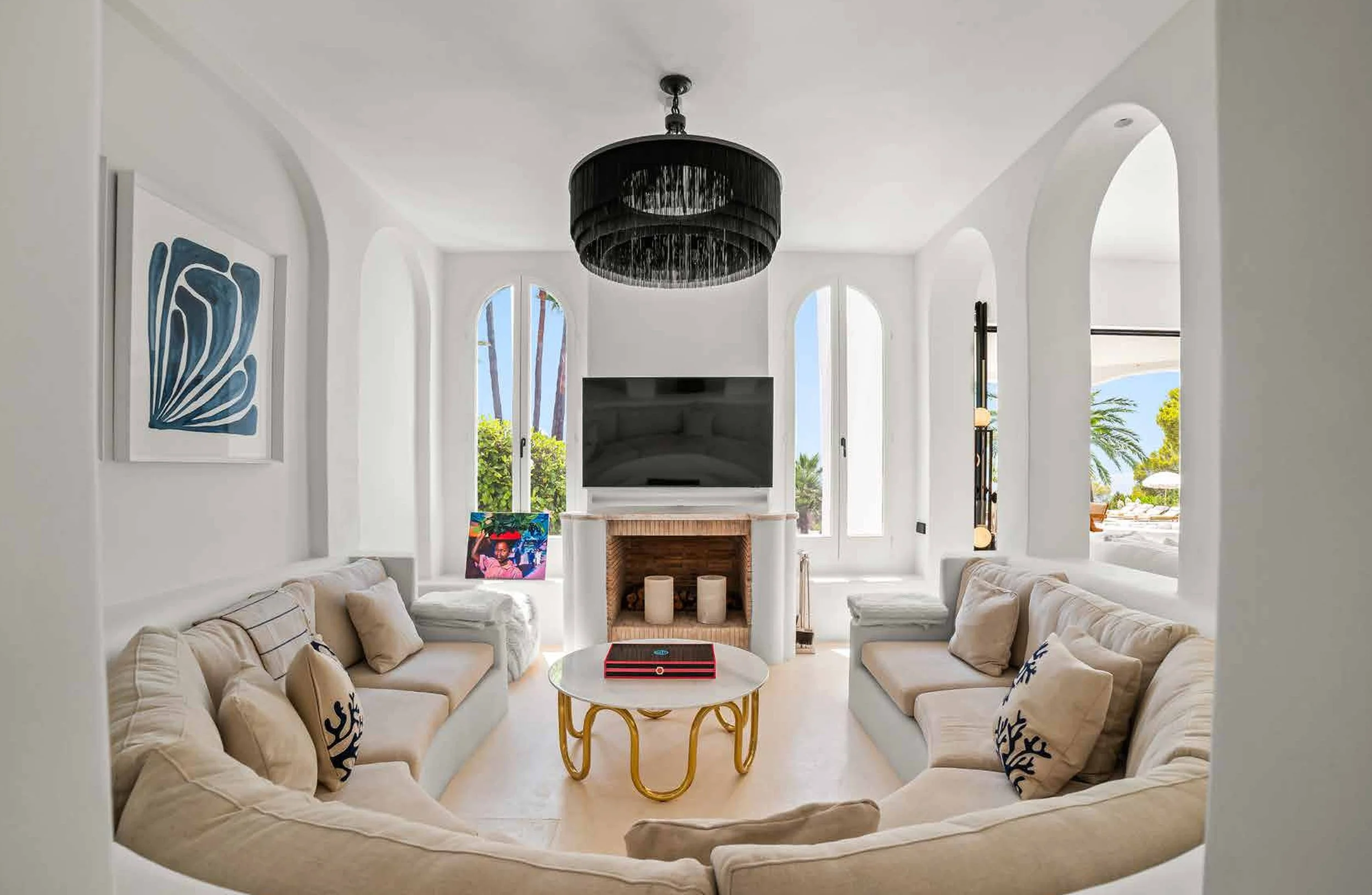 Bright living room with white walls, beige sofas, black chandelier, wall art, and large windows showing palm trees and outdoor scenery.