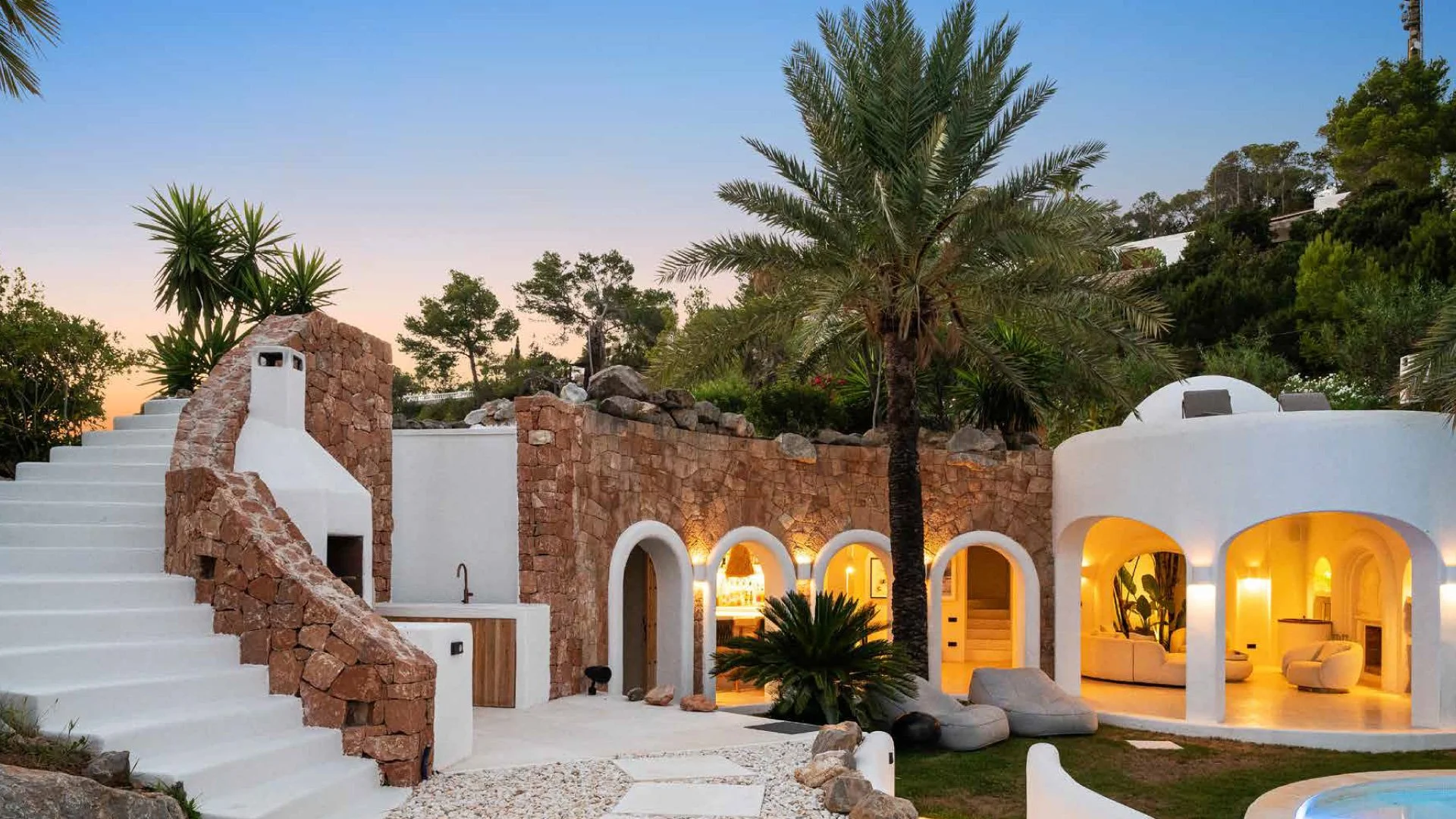 A modern, Mediterranean-style house with white walls and arched doorways, set among lush tropical plants and trees, with a sunset sky in the background.