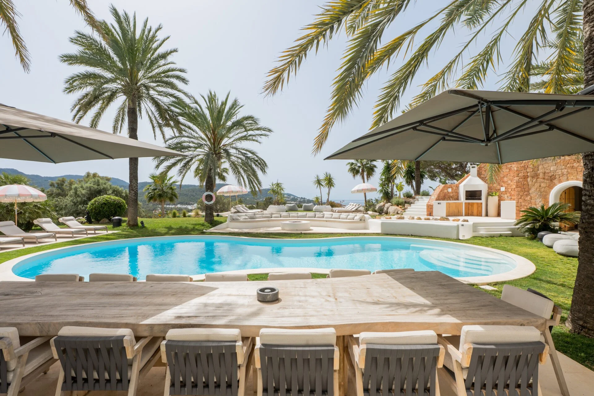 Ibiza villa outdoor large dining table with curved Swimming pool in a backyard with patio furniture, large umbrellas, palm trees, and a brick wall with an outdoor fireplace, overlooking a scenic view of mountains.