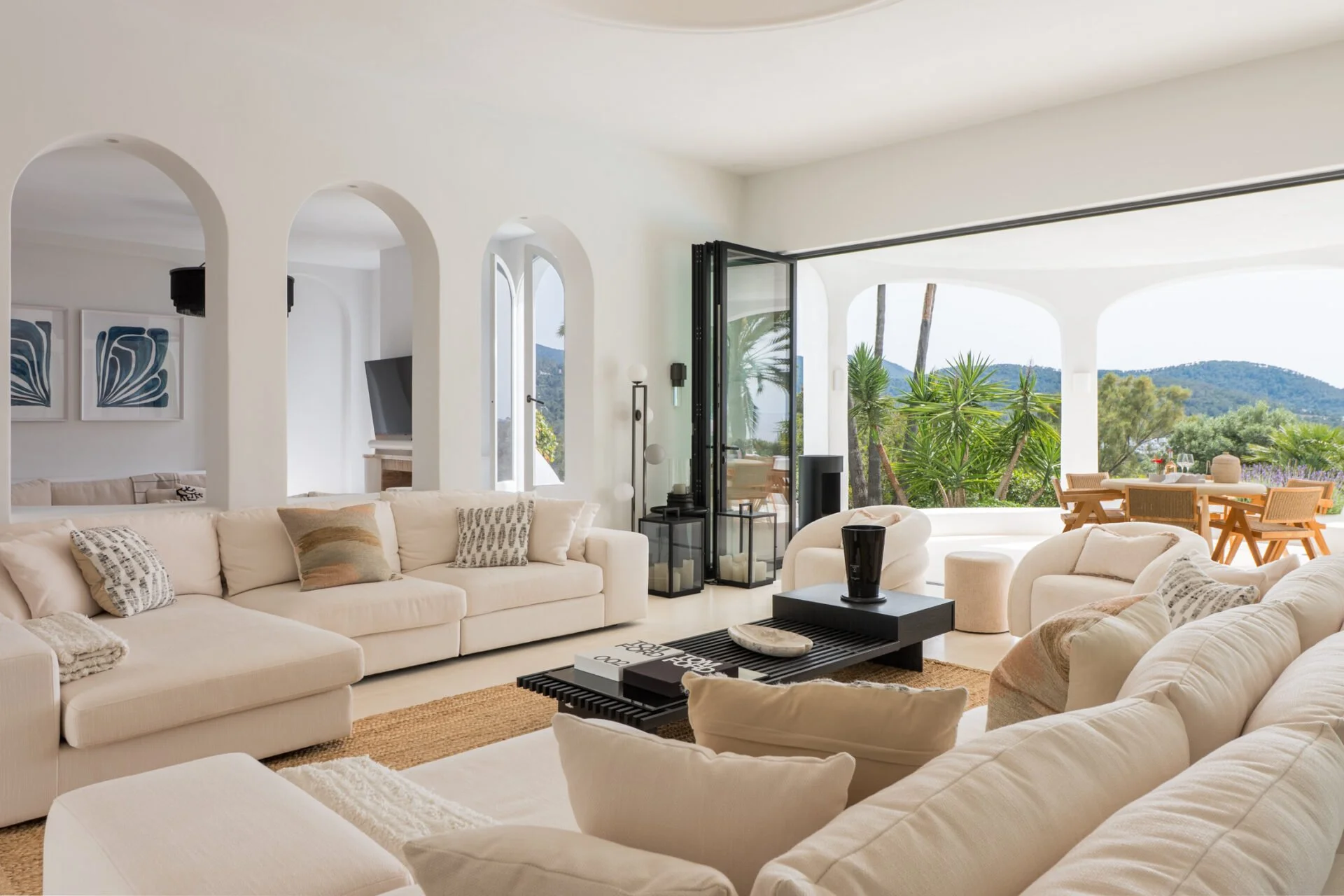 Bright living room with white sofas and neutral decor, large open sliding glass door leading to an outdoor patio with greenery and mountain view.