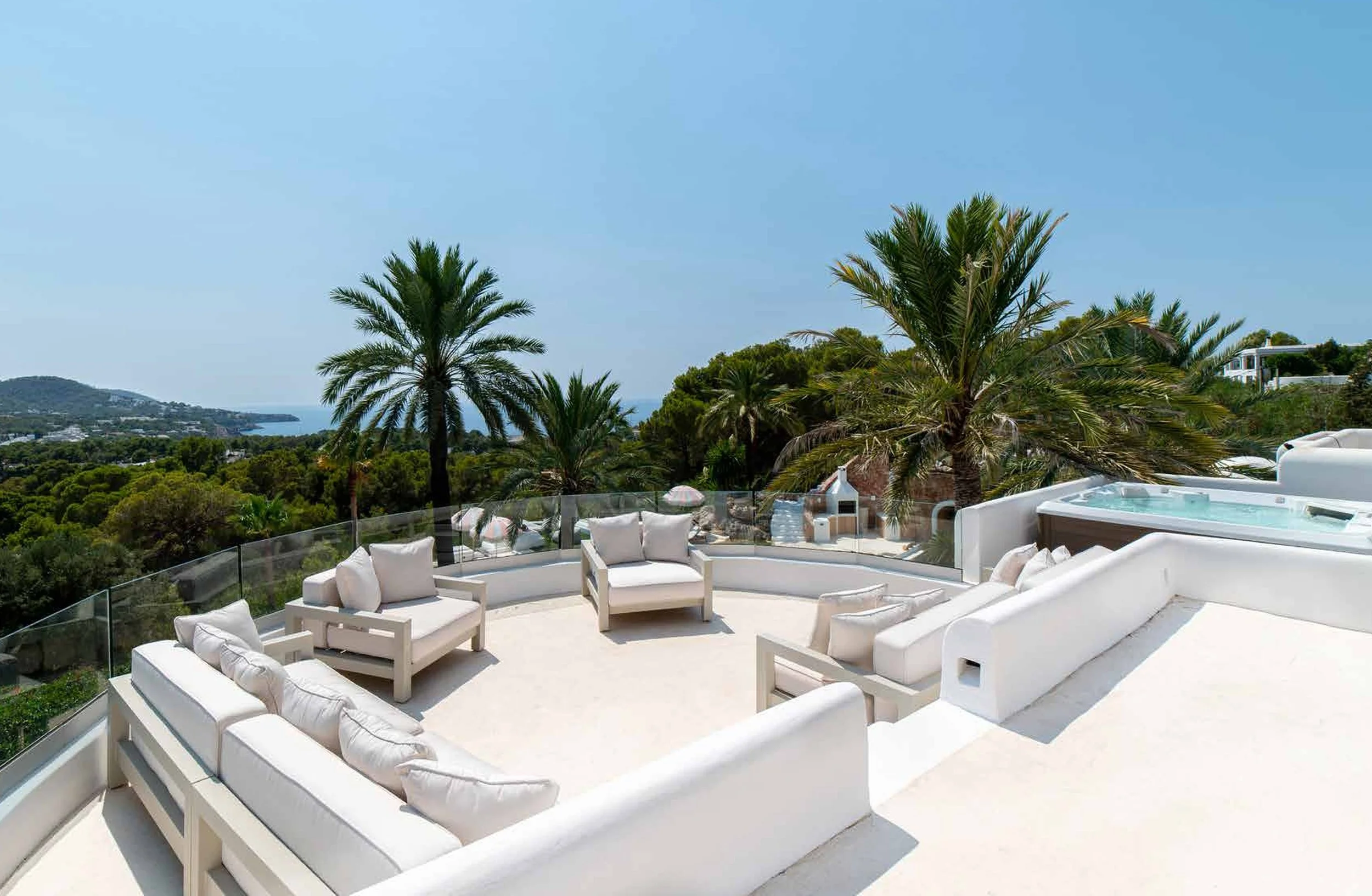 Luxury outdoor patio with white seating, palm trees, and a view of the ocean and hills in the distance.