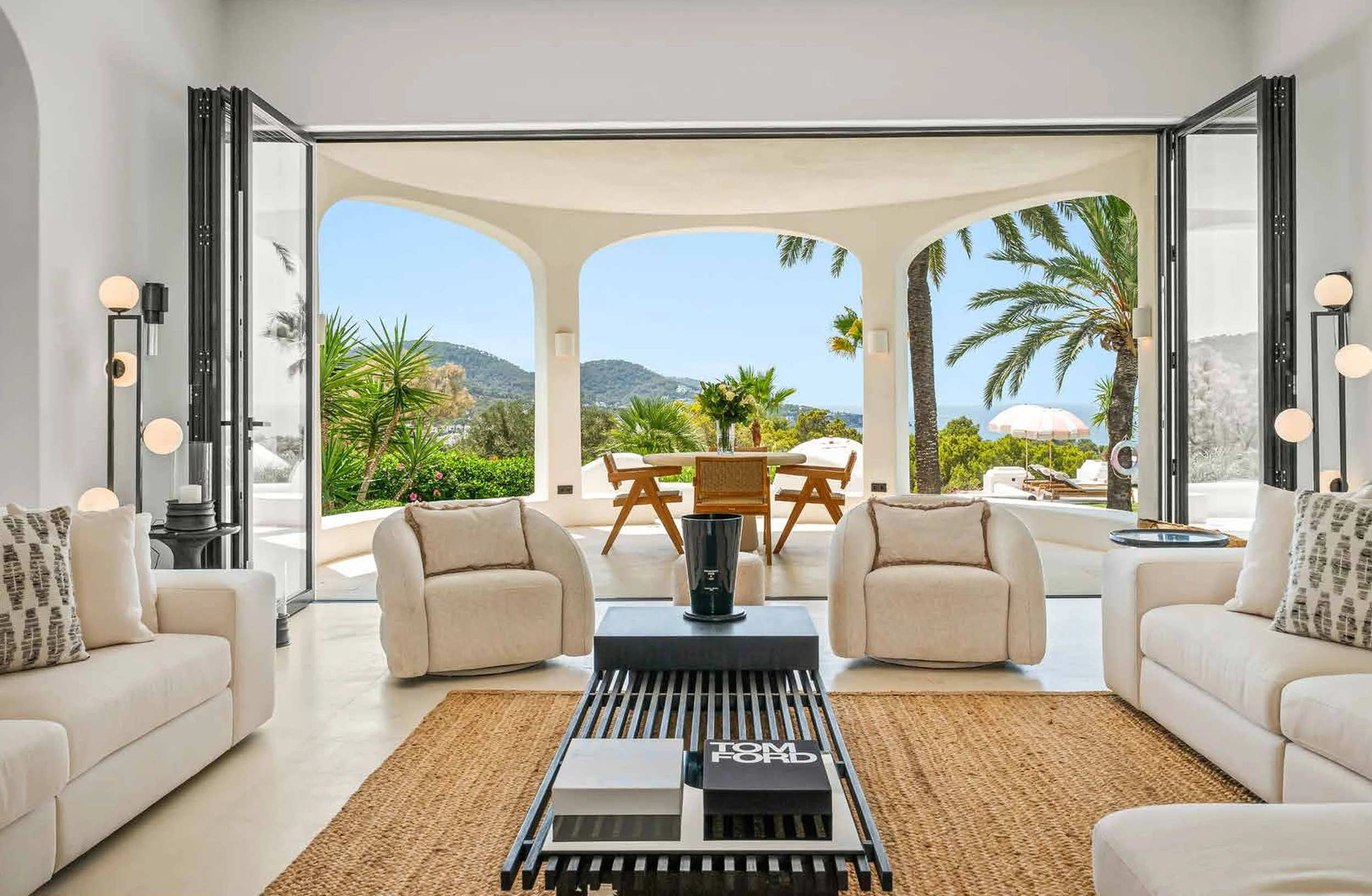 Luxurious living room with open sliding doors leading to a terrace with ocean view, outdoor dining table, lush greenery, and palm trees.