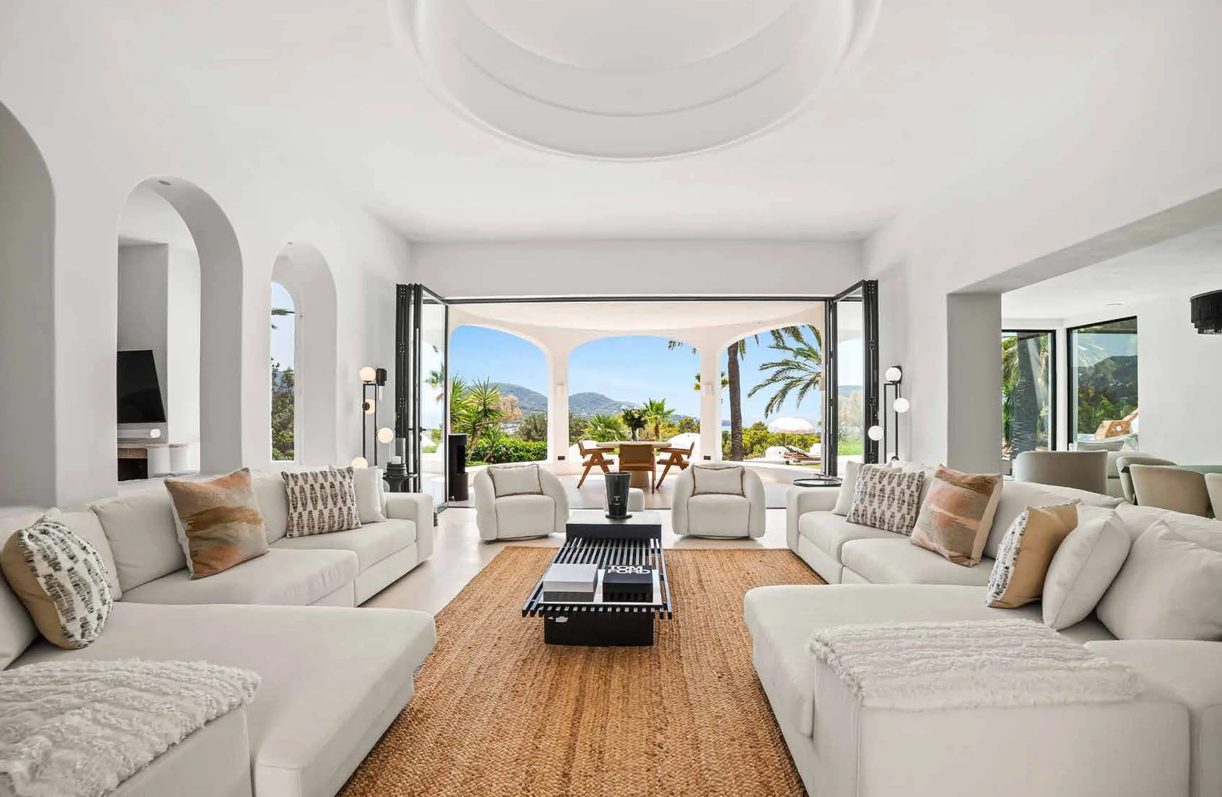 Bright, spacious living room with white sofas and armchairs, a central black coffee table, and scenic view of palm trees and mountains through open French doors leading to a patio
