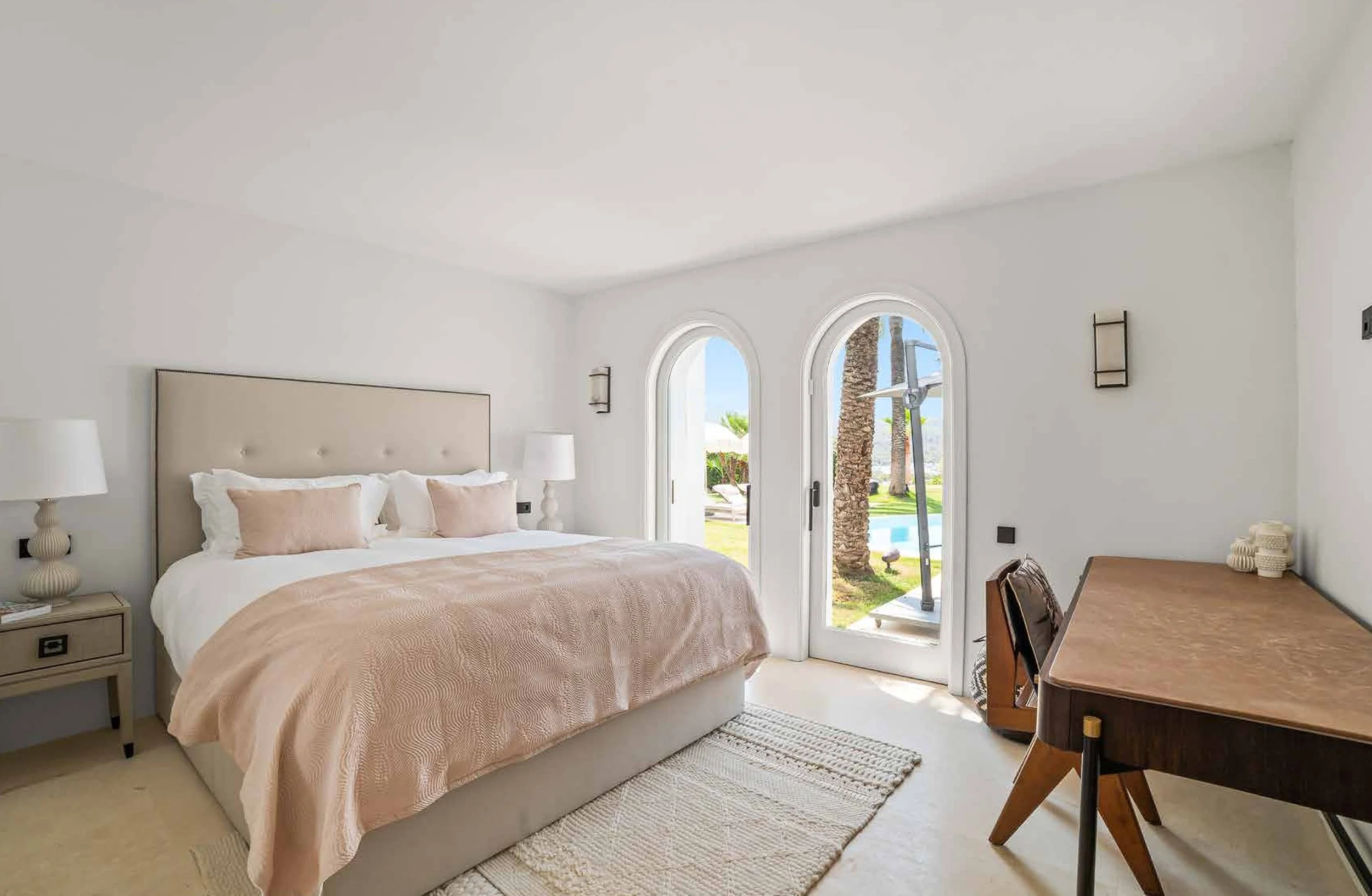 A bright, modern bedroom with a large bed, beige headboard, and light pink bedding, next to a window showing a backyard with palm trees and a swimming pool, complemented by a wooden desk and chairs.