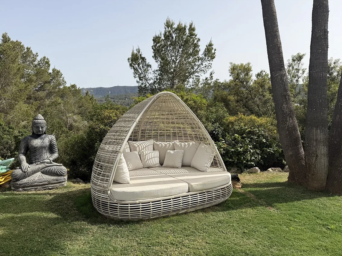 A modern outdoor rattan canopy daybed in an ibiza villa with white cushions on a lawn, surrounded by trees and bushes, with a Buddha statue nearby. Ibiza style garden.