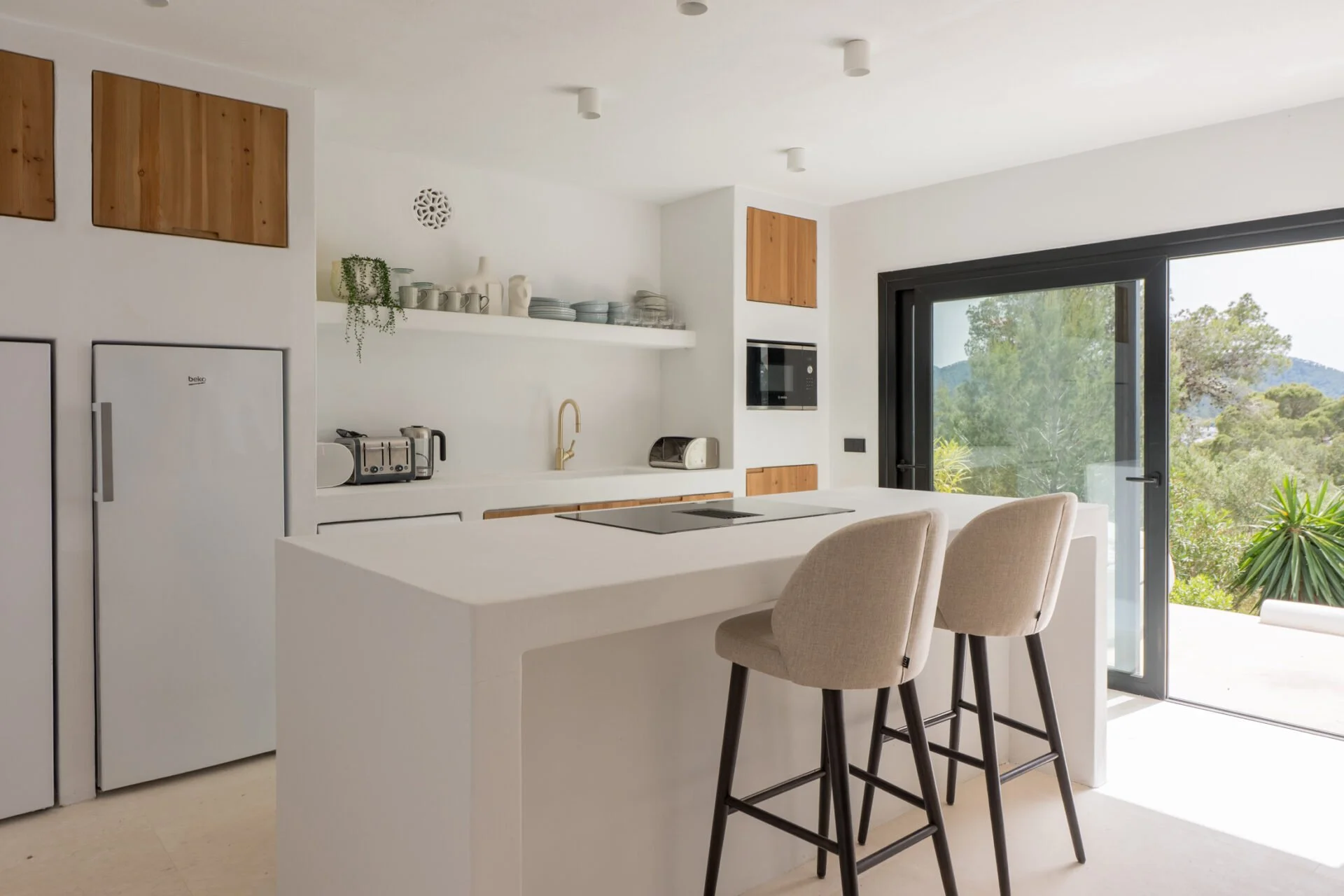 Modern kitchen with white cabinets, a large island, beige upholstered chairs, open shelving with dishes, a black-framed sliding glass door, and outdoor greenery visible through the window.