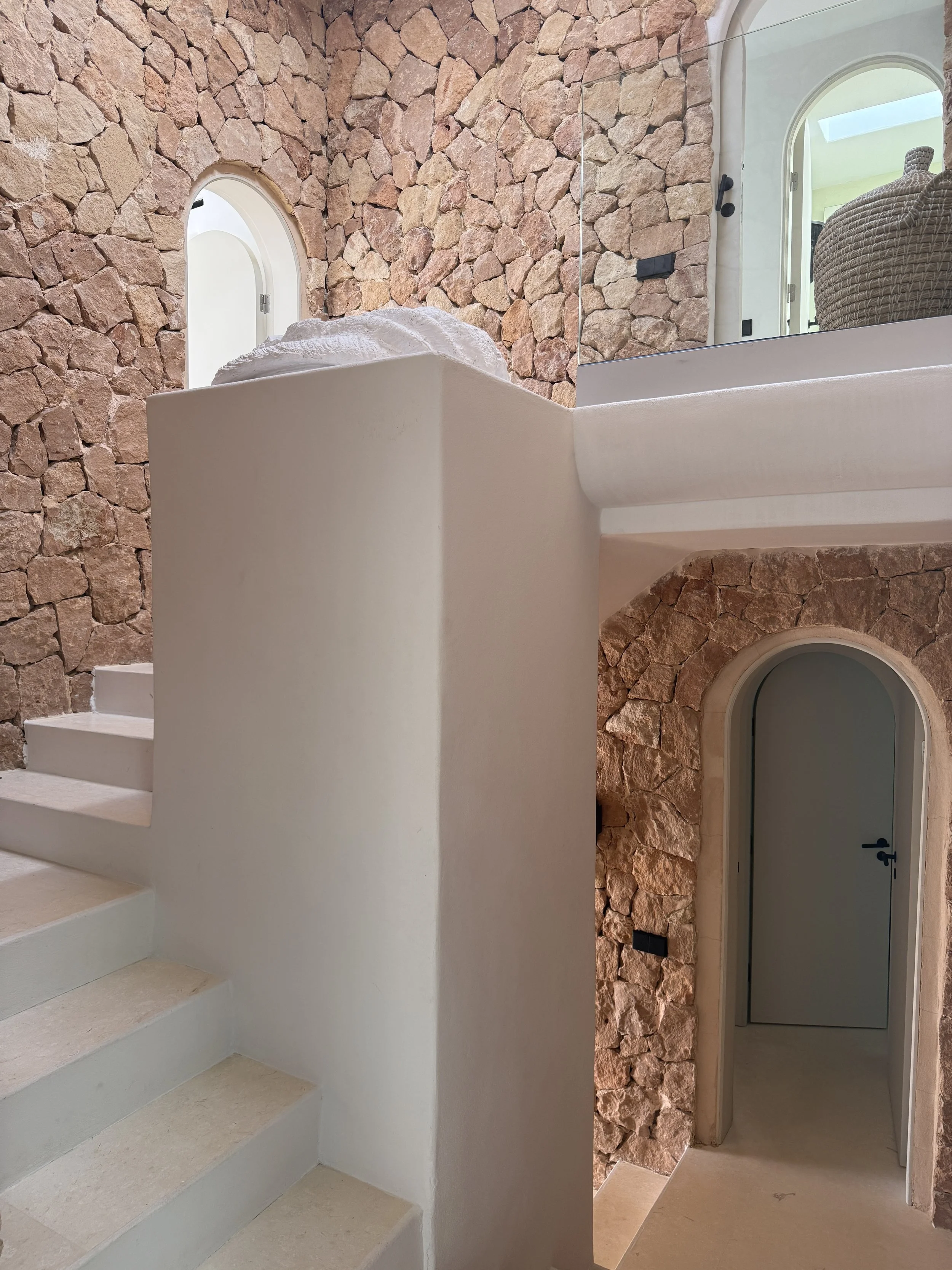 Interior of a house featuring a stone wall with arched doorways and stairs, and a curved staircase railing.