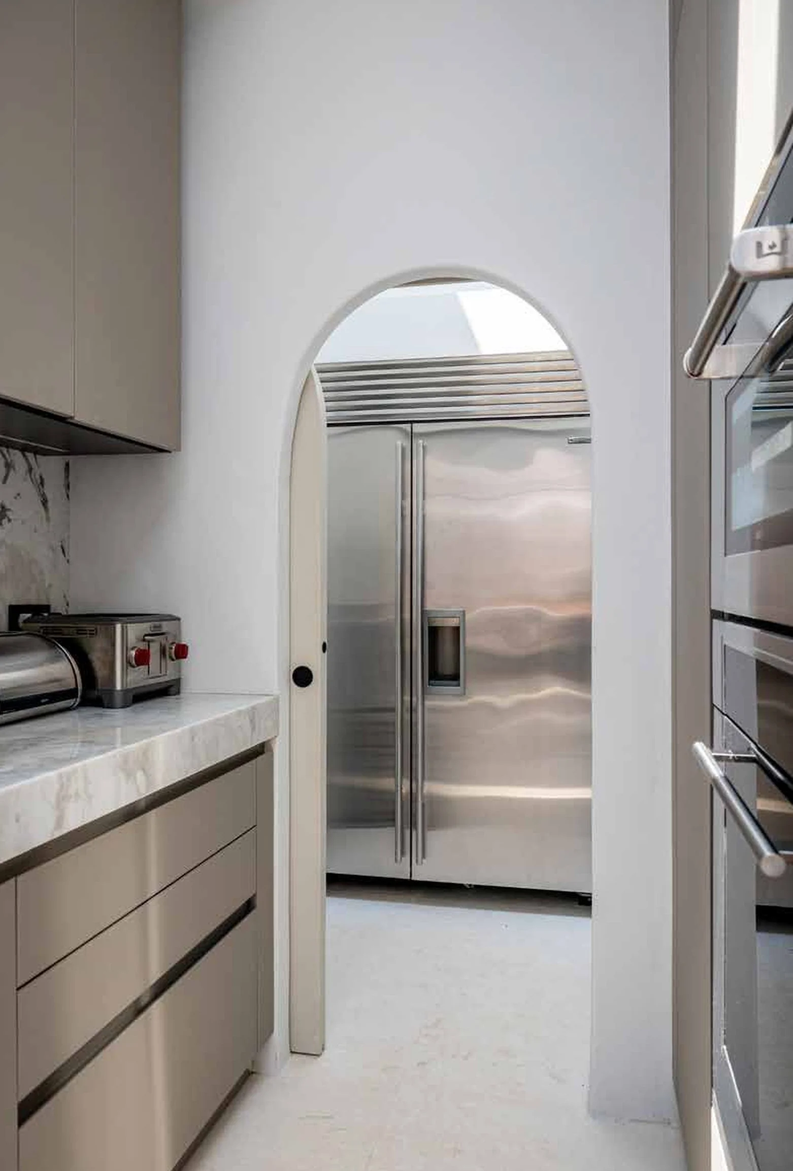 Modern kitchen with white walls, beige cabinets, and stainless steel appliances including a large refrigerator and an oven. A marble countertop is visible on the left side.
