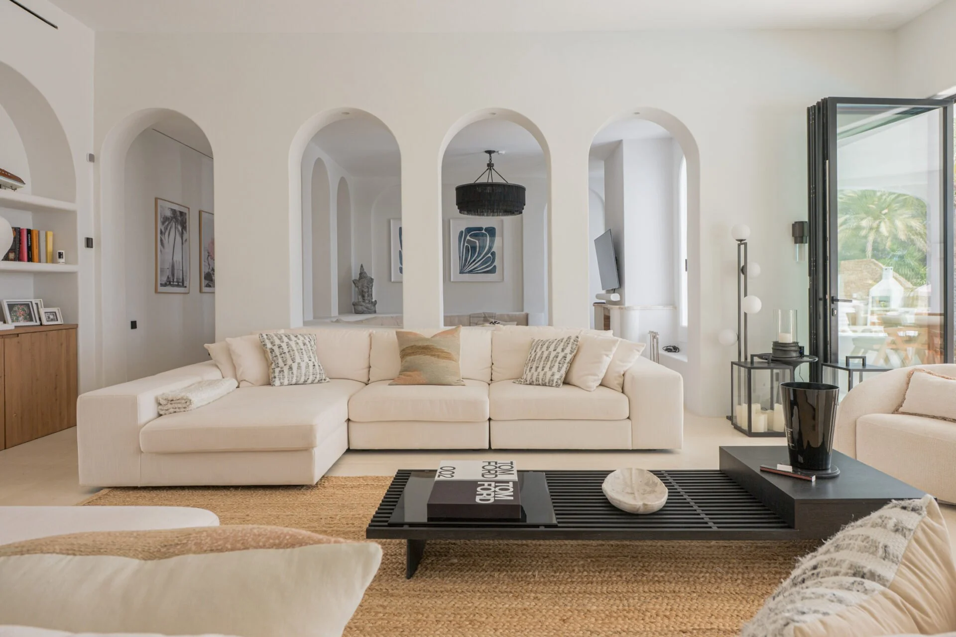A bright, modern living room with a white sectional sofa adorned with decorative pillows. There is a black coffee table with books and a decorative object, set on a natural fiber rug. Behind the sofa, an open wall features arched openings, art prints