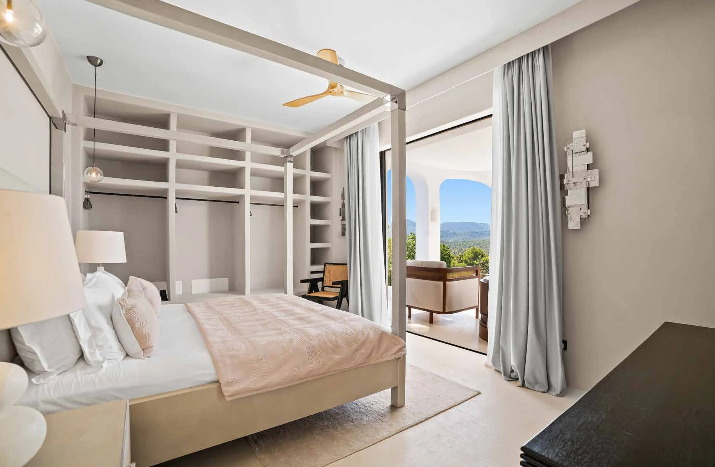 A bedroom with a four-poster bed, white bedding, and bedside lamps. There is a balcony with outdoor seating and a view of mountains and trees in the background.