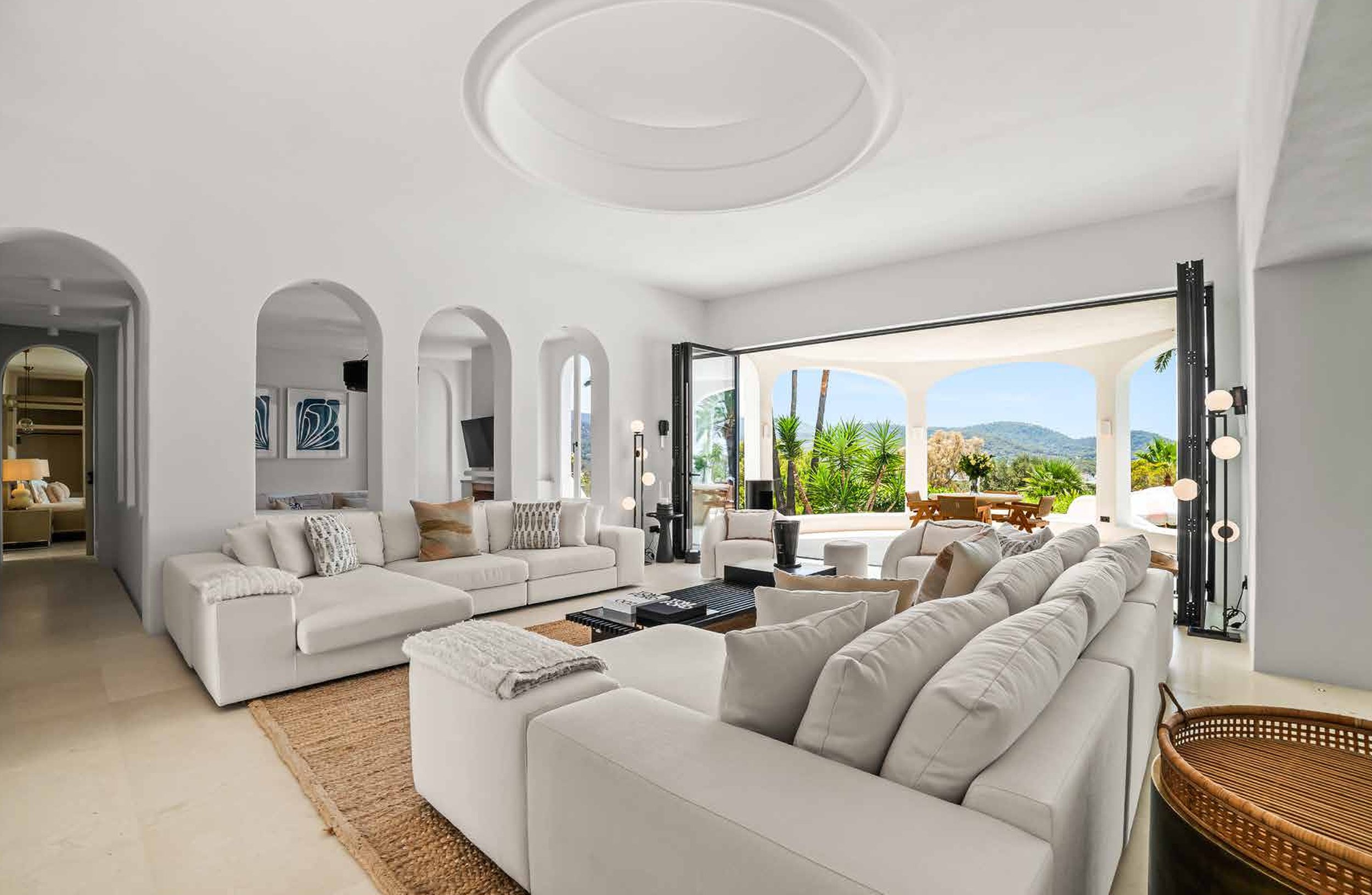 Bright, spacious living room with white sofas, open door leading to an outdoor patio with palm trees, sea view, and mountain scenery in the background.