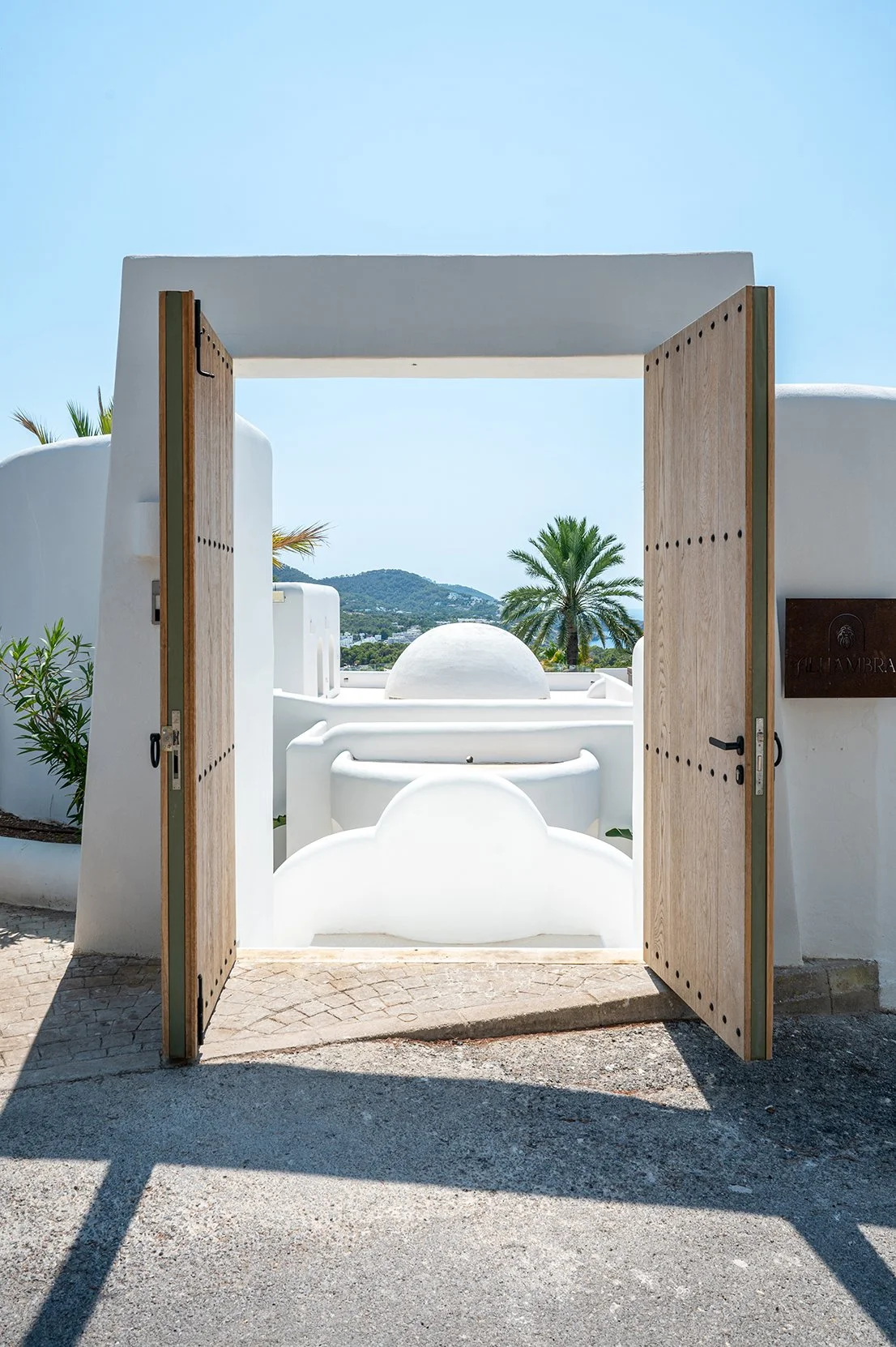 Open wooden door leading to white Mediterranean-style buildings with a background of palm trees and hills under a clear blue sky.