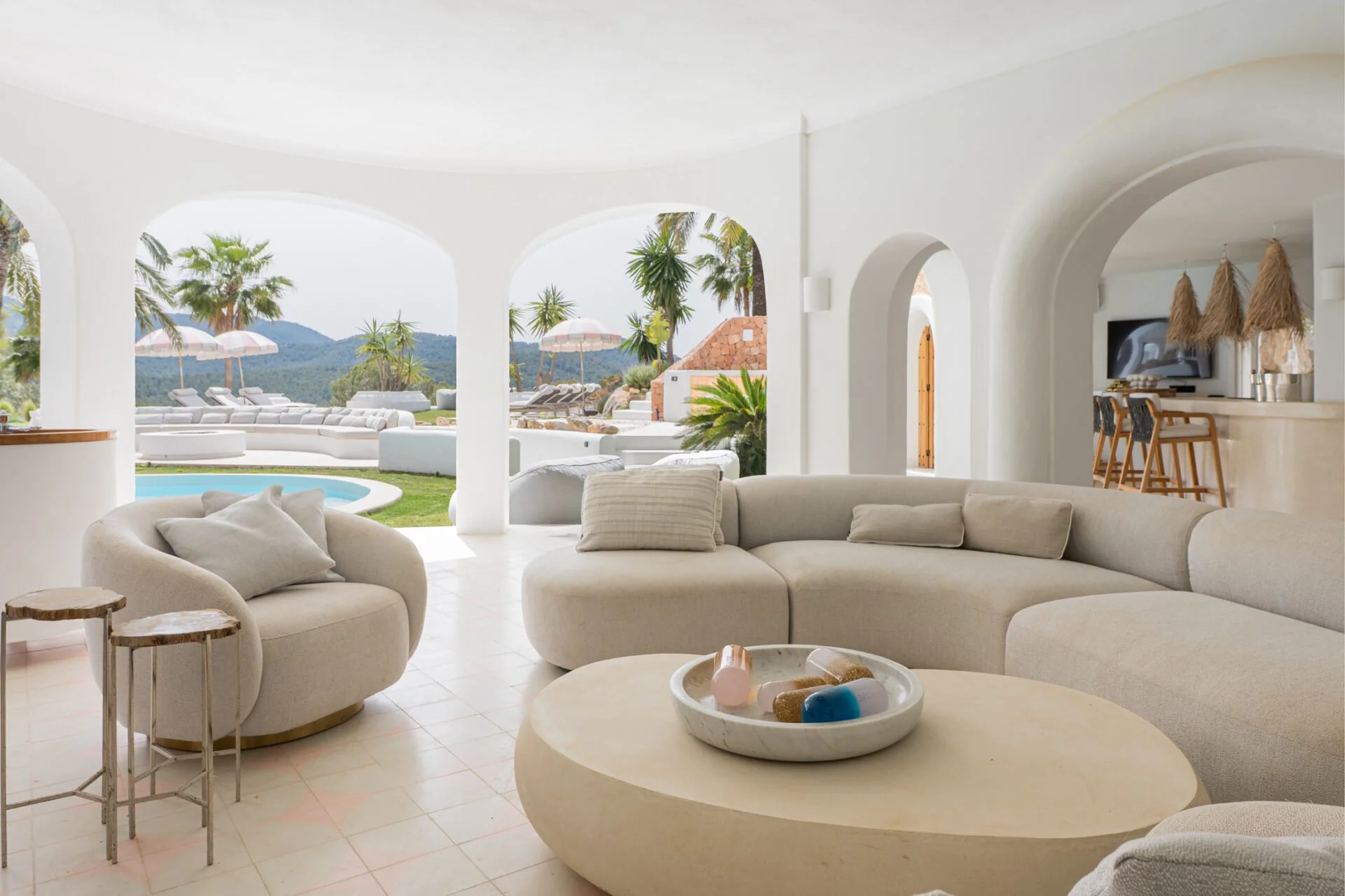 Interior of a bright, white, Mediterranean-style living room with curved archways opening to a poolside patio with lounge chairs and umbrellas, surrounded by palm trees and mountains.