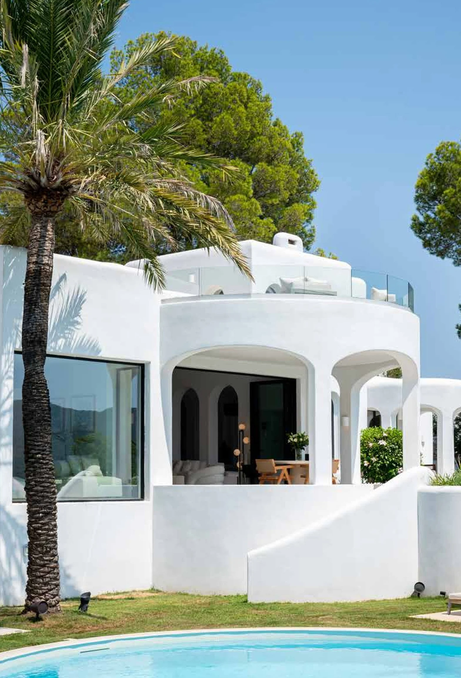 White modern house with curved architecture, large glass windows, surrounded by green trees, with a swimming pool in the foreground.