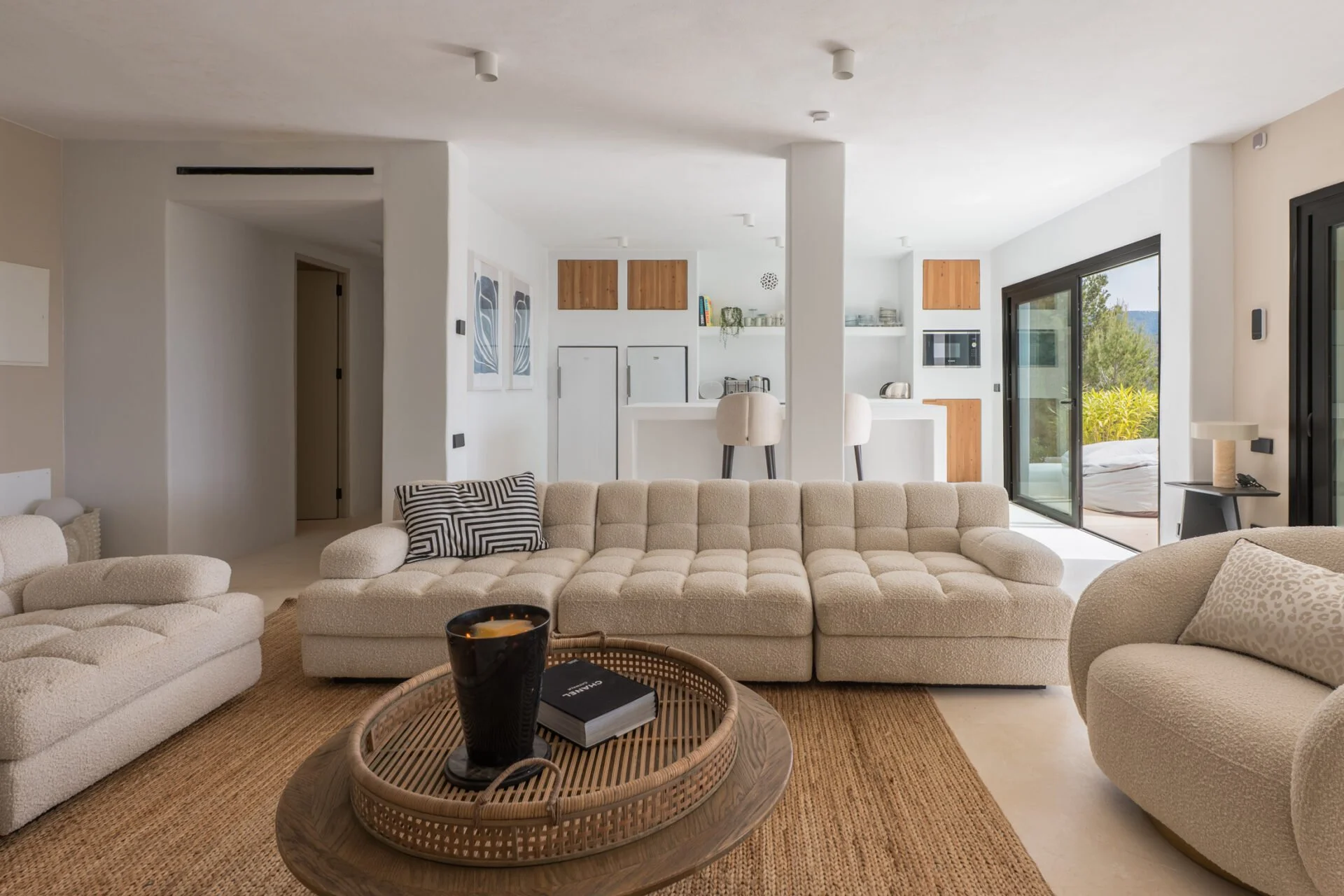 Living room with beige sectional sofa, coffee table with candle and book, and sliding glass door leading to an outdoor patio with green trees.
