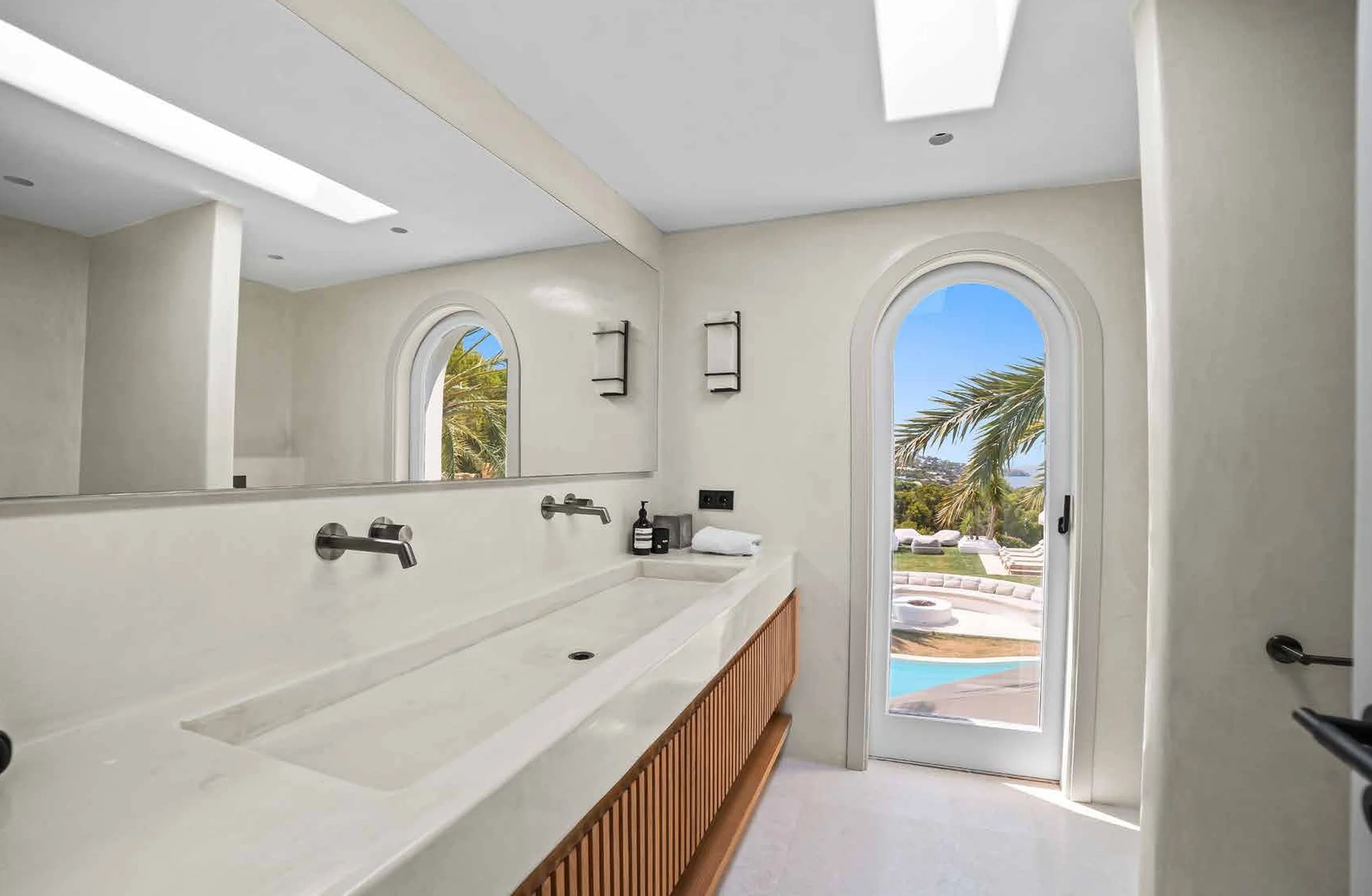 Modern bathroom with white wall, large mirror, dual wall-mounted faucets over a rectangular sink, and a door with a view of an outdoor pool and palm trees.