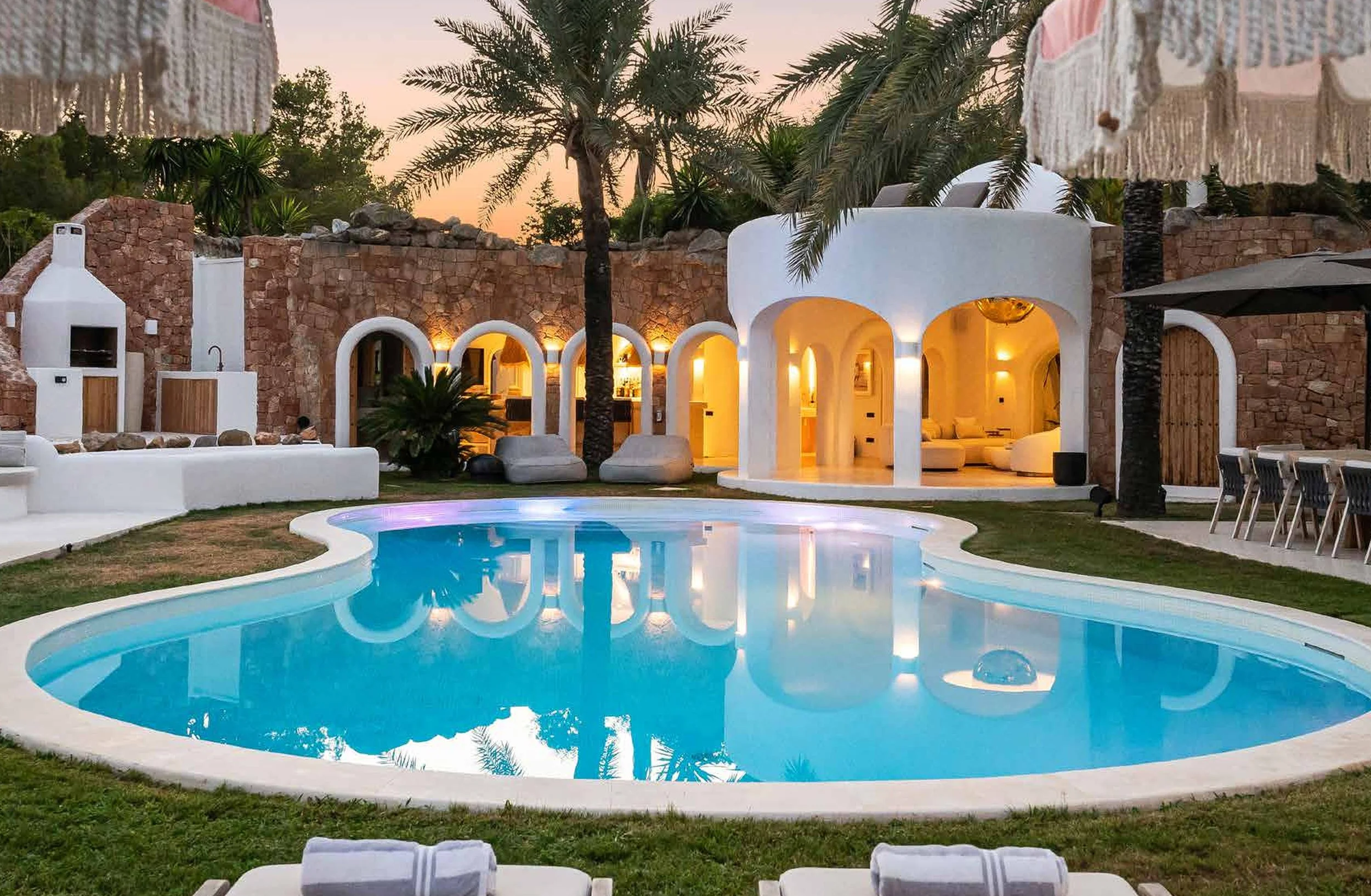 A luxurious backyard with a kidney-shaped swimming pool, surrounded by green grass, palm trees, and outdoor seating areas. There are outdoor fireplaces and a white stucco house with arched doorways, all illuminated by warm lighting at dusk.