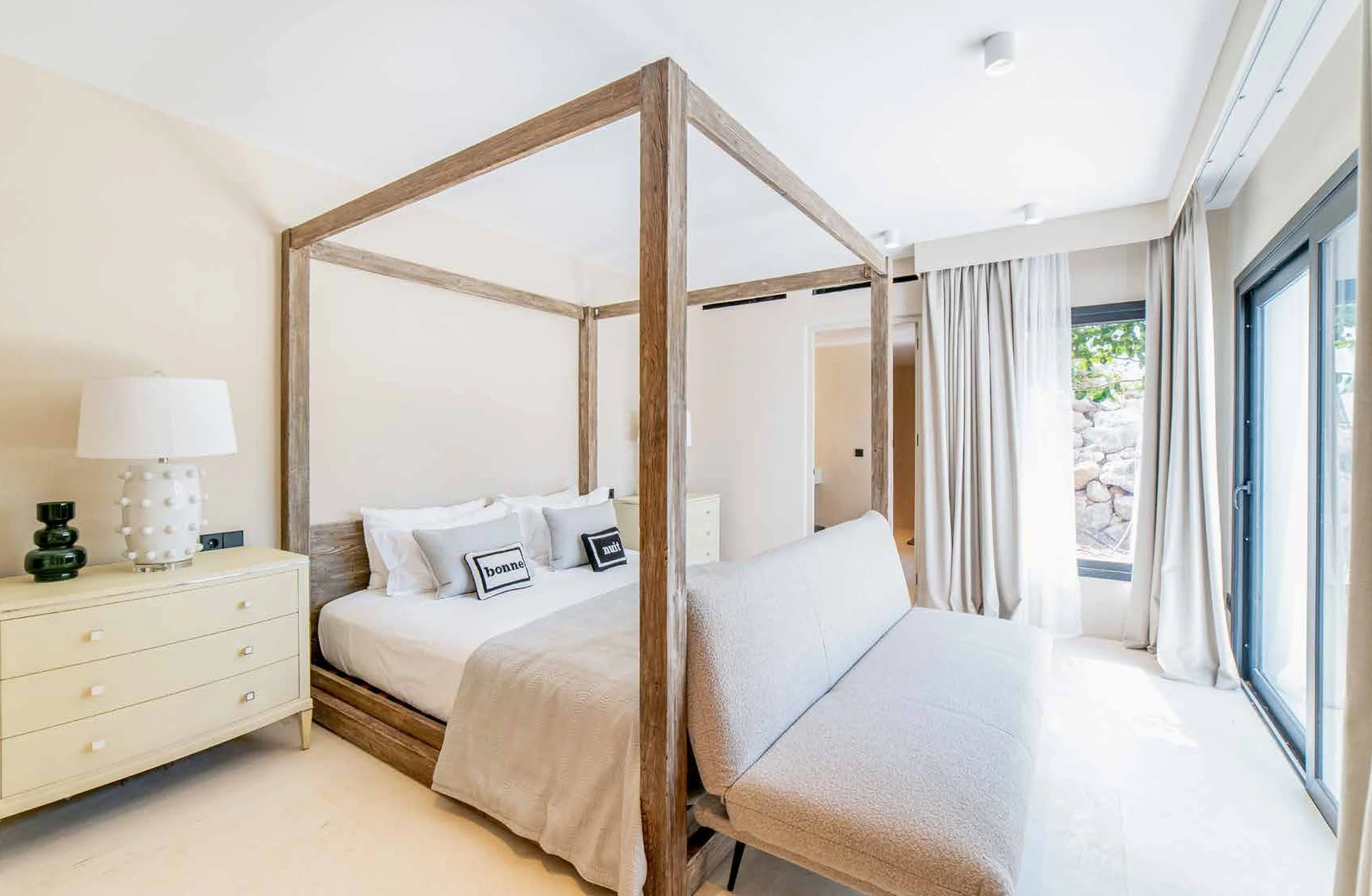 A bright bedroom with a large bed, four pillows with words 'bonne nuit' on them, a cream dresser with a white lamp and decorative black objects, a beige bench at the foot of the bed, large windows with white curtains, and a small outdoor area with rocks visible outside.
