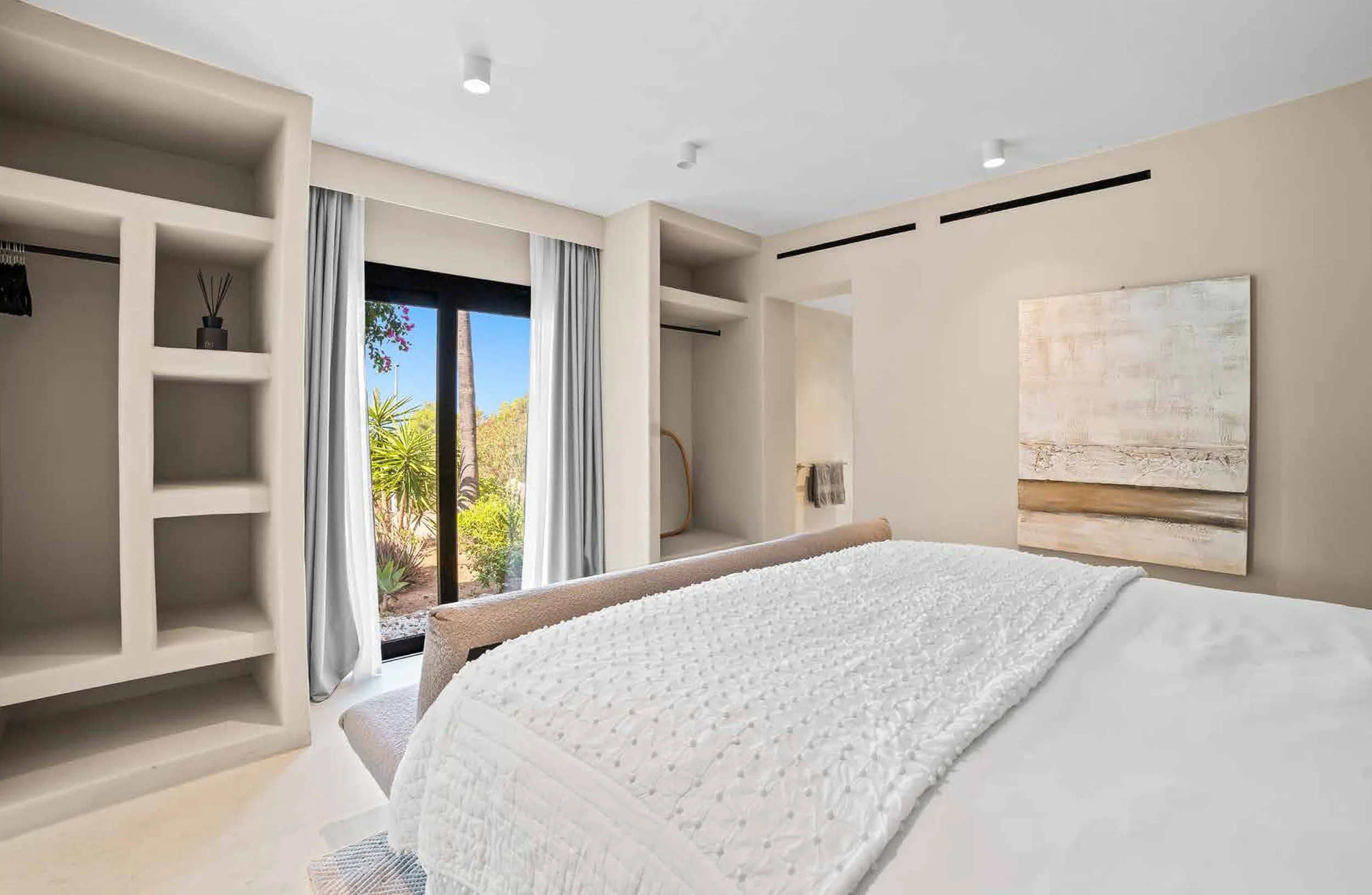 Modern bedroom with white bedding, abstract wall art, open closet space, and sliding glass door leading outside to greenery.