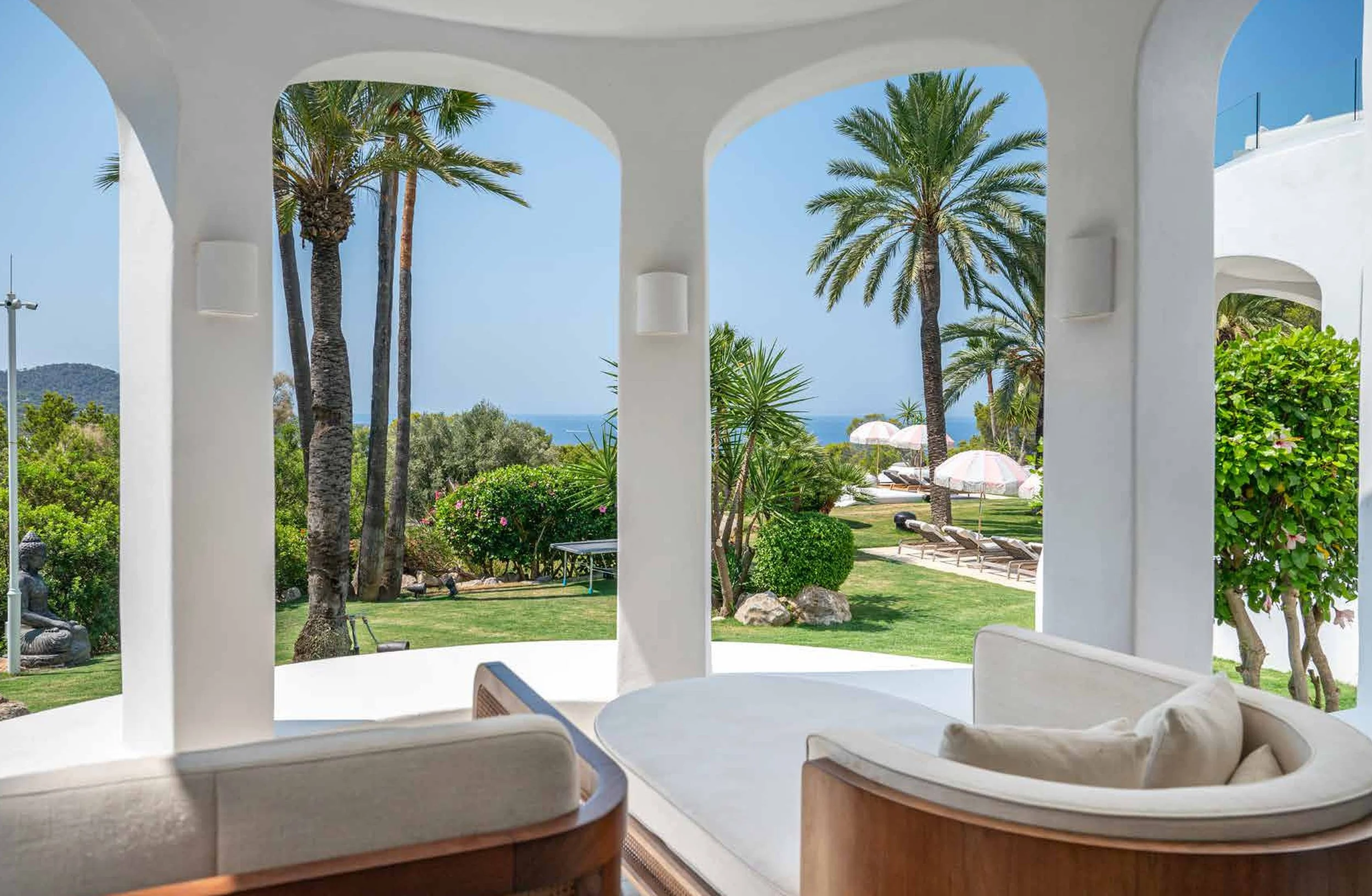 View from a white curved porch with white walls looking out onto a lush tropical garden with palm trees, shrubs, and flowers, and lounge chairs with pink umbrellas near the sea in the background.