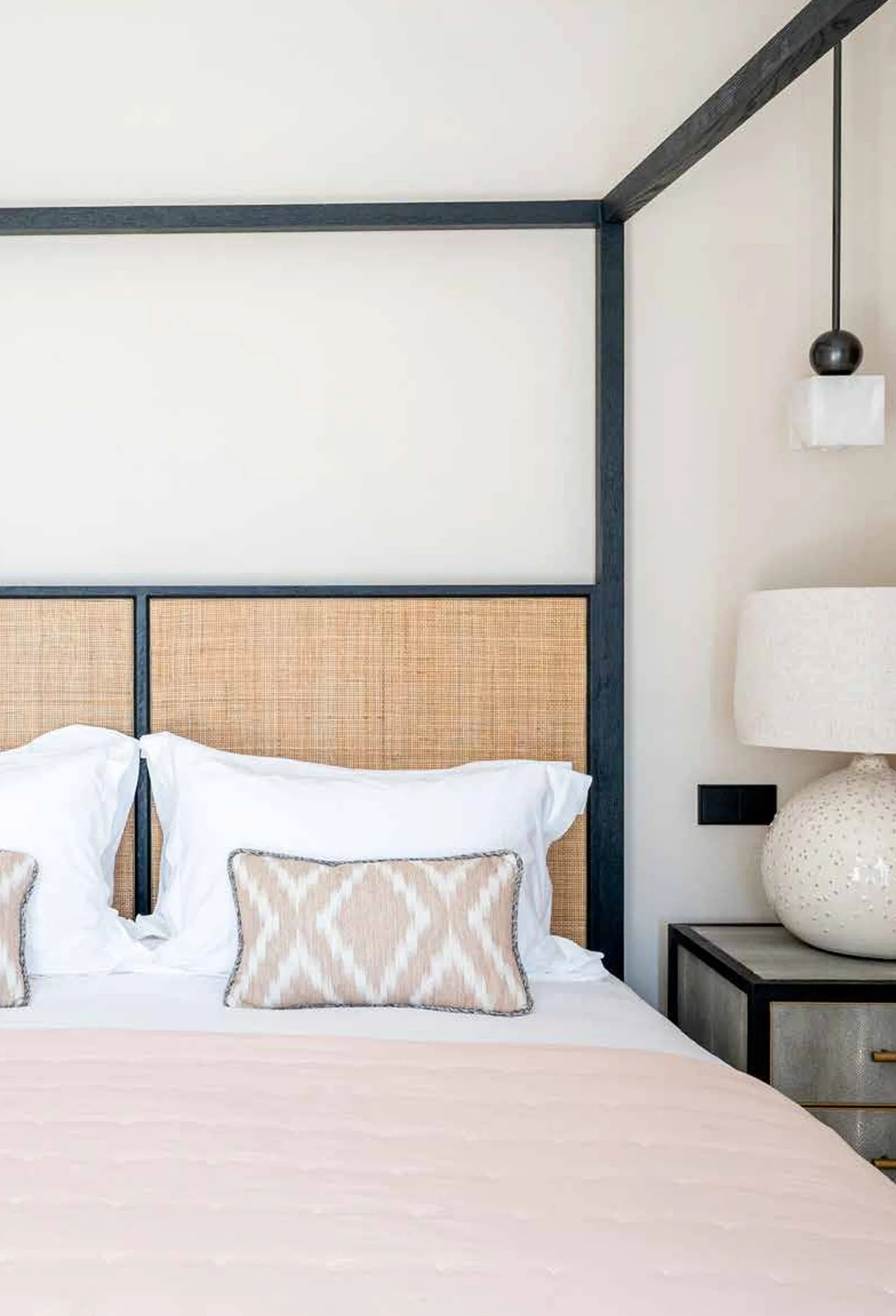 A bedroom with a bed that has white sheets, beige decorative pillows, and a woven headboard framed with black wood. To the right, there is a bedside table with a large white textured lamp and a black wall socket or switch. A ceiling pendant light wit