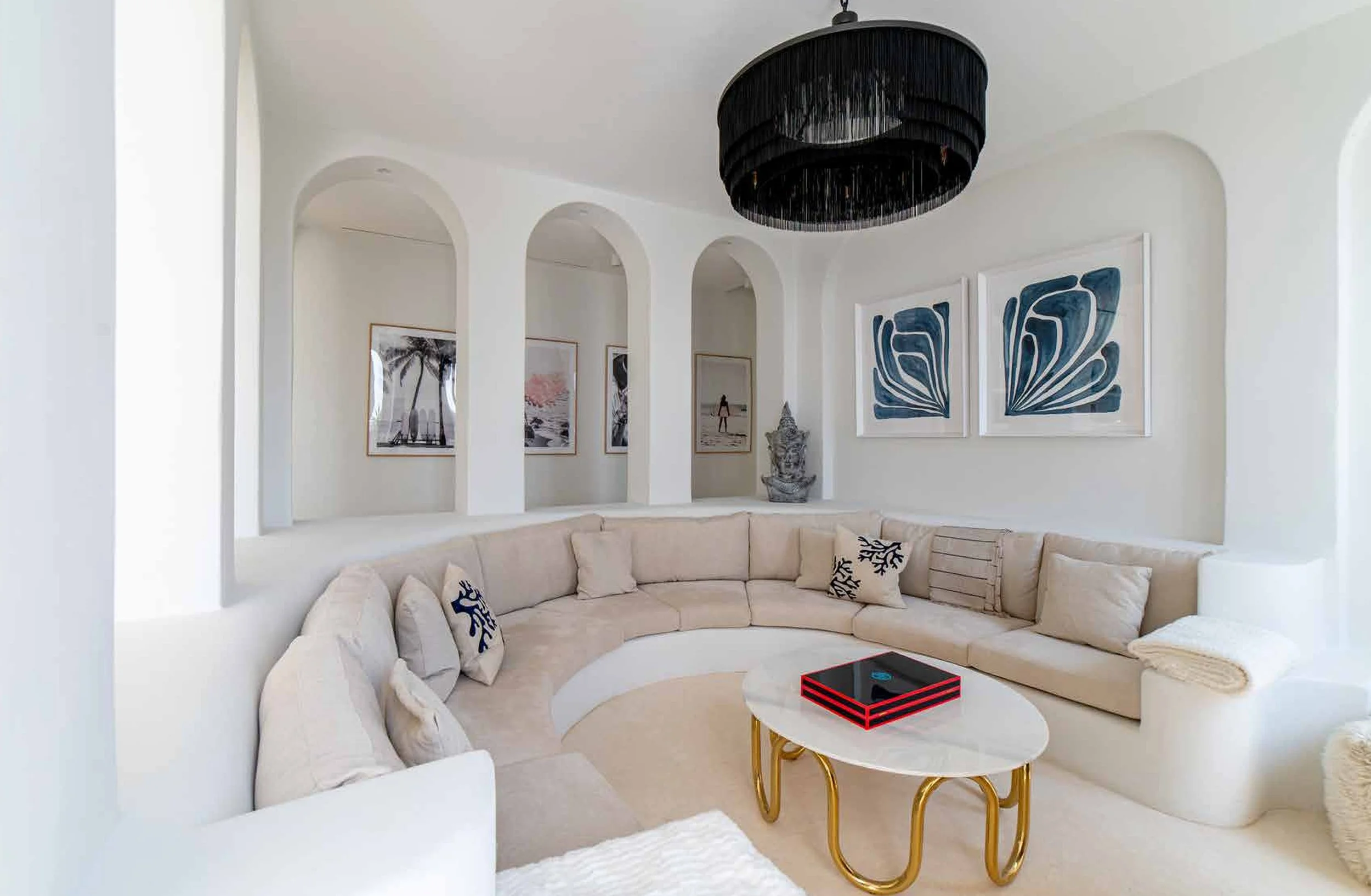 Living room with white curved sectional sofa, decorative throw pillows, a marble coffee table with a red and black box, white walls with blue abstract art, black circular chandelier, framed pictures and statues in the background.