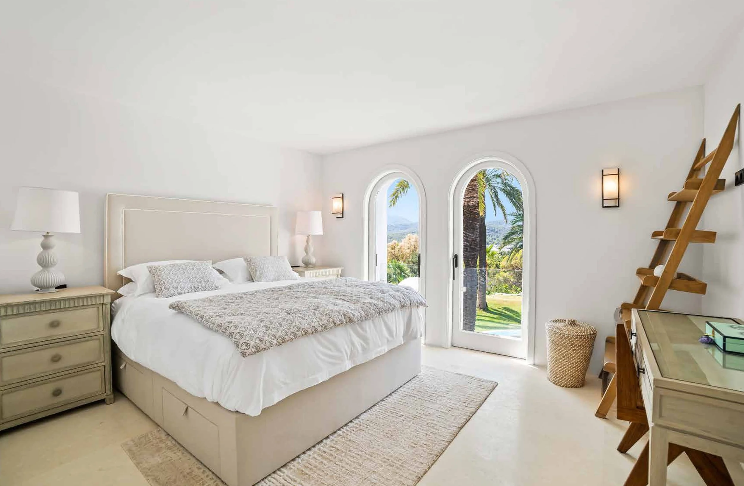 Bright bedroom with white walls and furniture, large bed with patterned bedding, two nightstands with lamps, sliding glass door with an outdoor view of trees and mountains, palm trees outside, wooden ladder, beige rug, wicker basket, and a dresser with green box