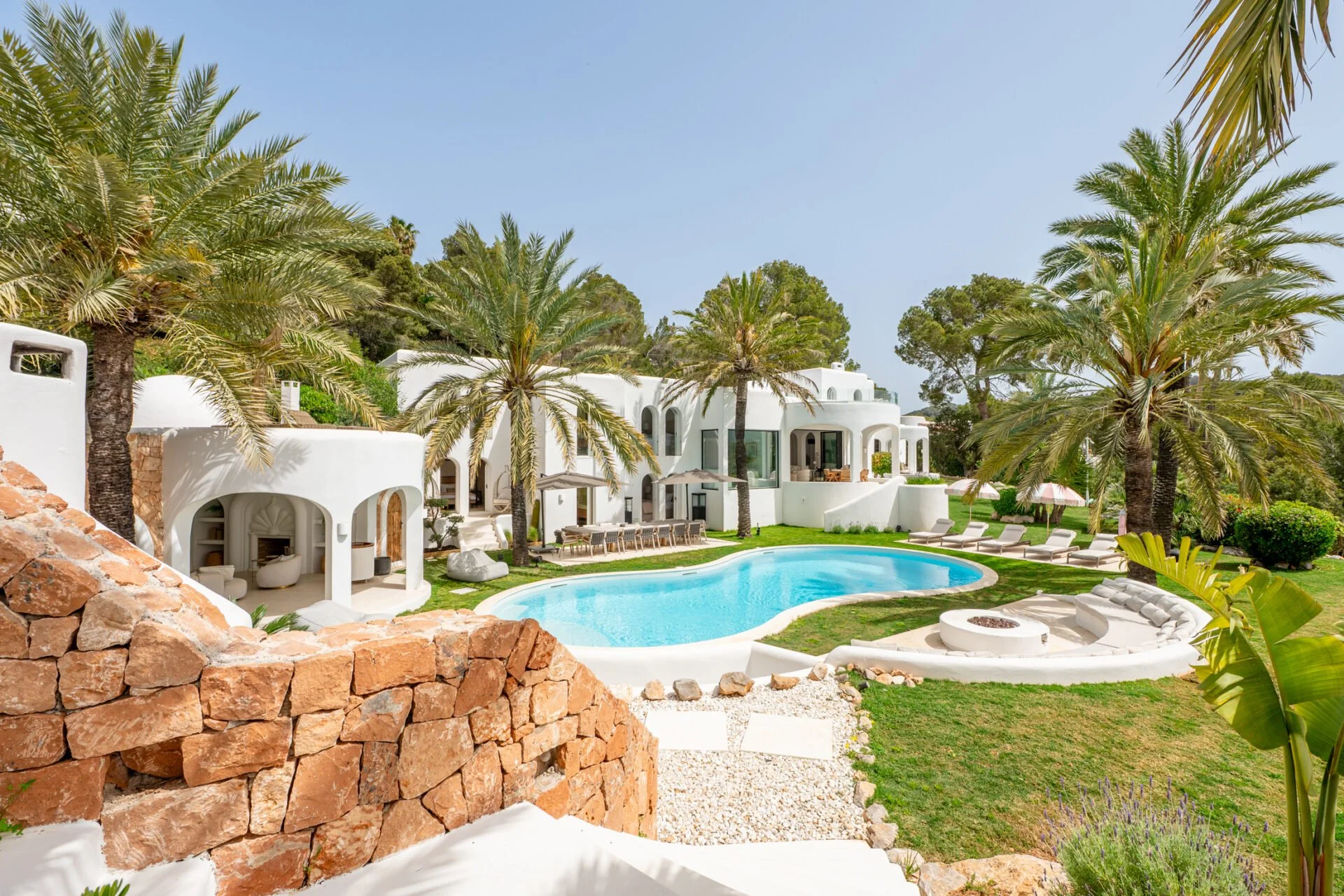 Beautiful white Mediterranean-style baleric villa with a swimming pool, surrounded by palm trees and lush greenery, under a clear blue sky.