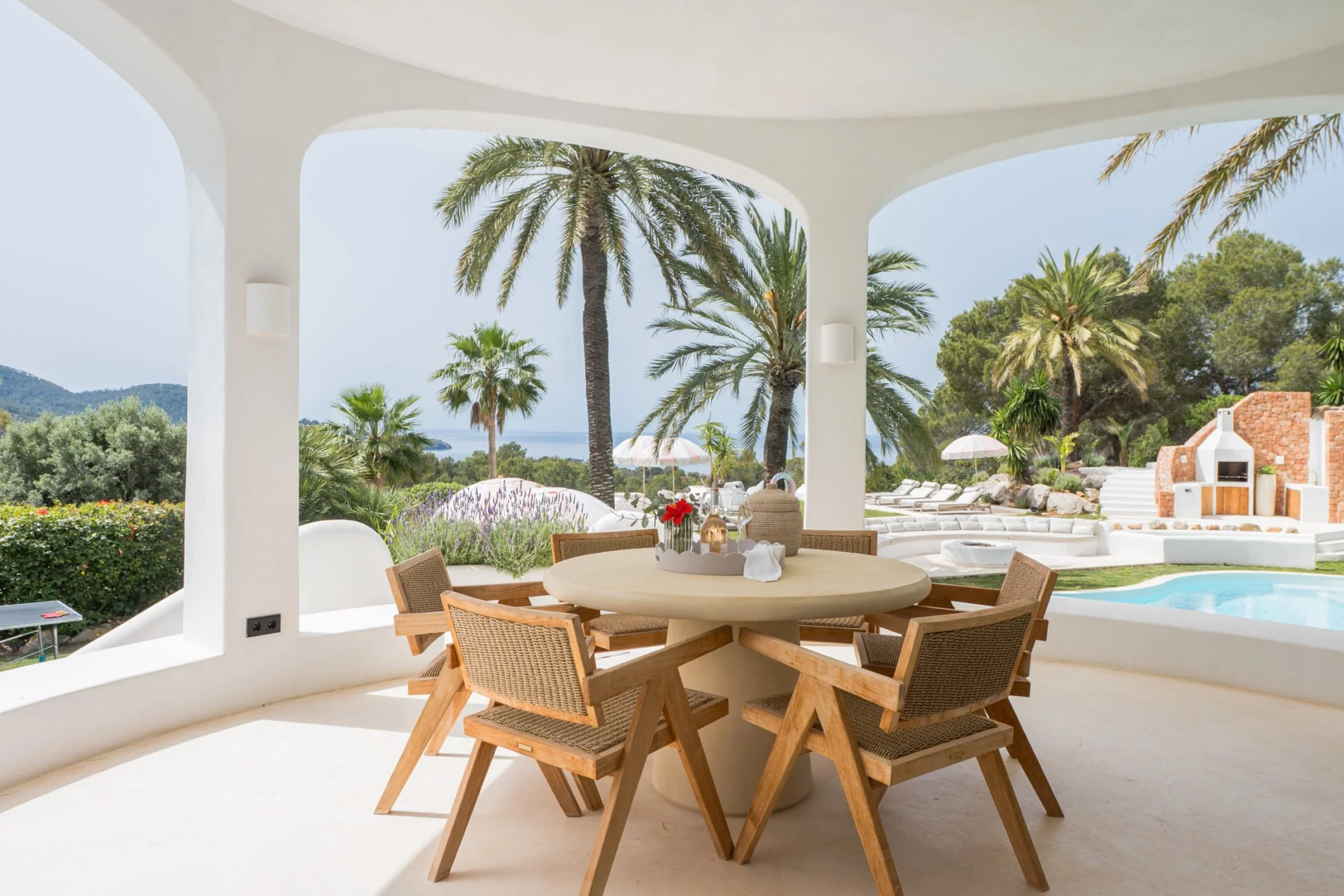 Outdoor patio with a round table and wooden chairs, overlooking a pool, palm trees, and a scenic landscape with hills in the background.