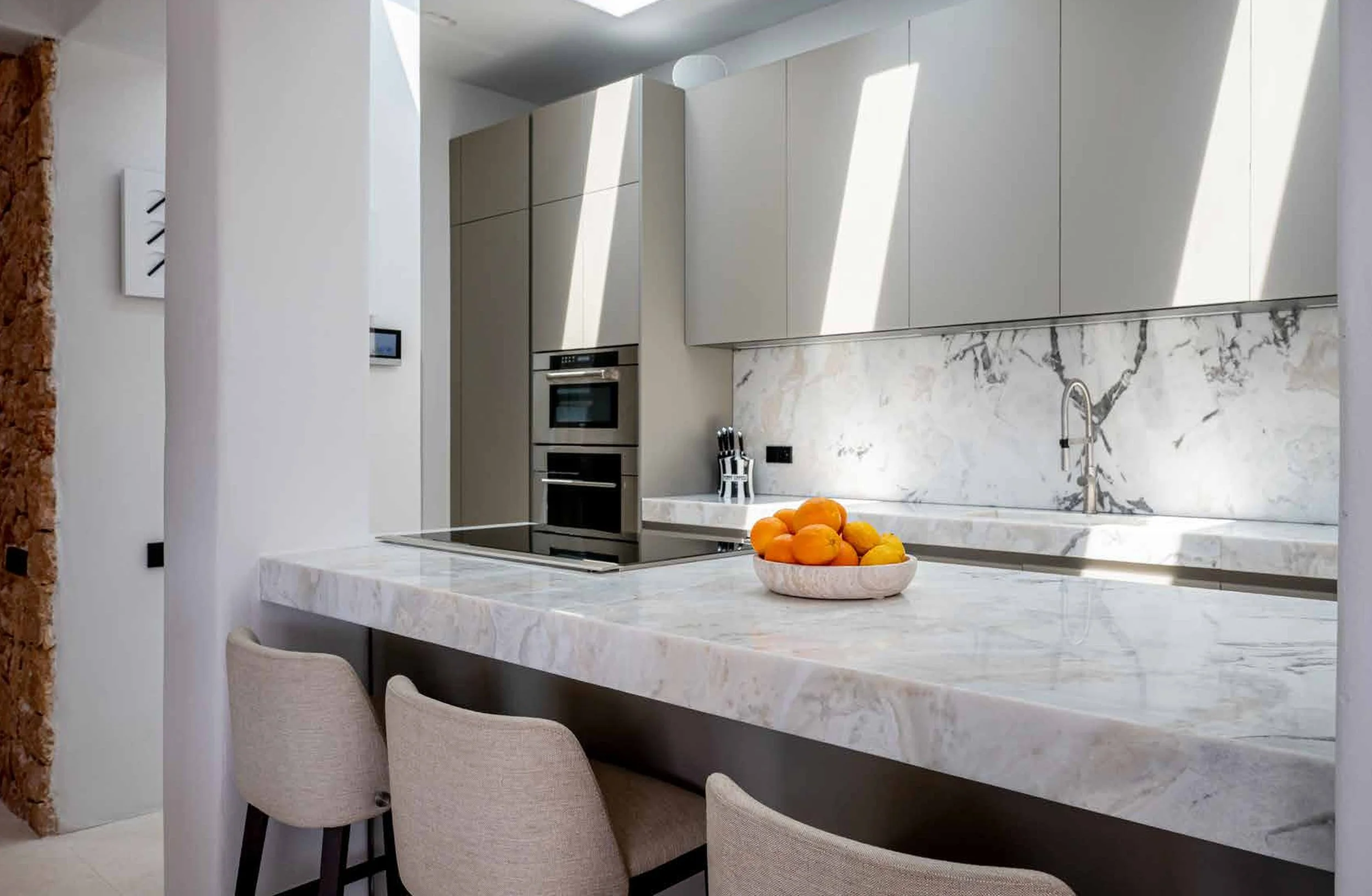 Modern kitchen with marble countertops, neutral cabinetry, and a bowl of oranges on the island. Built-in oven and microwave, faucet, and sunlight create a bright atmosphere.