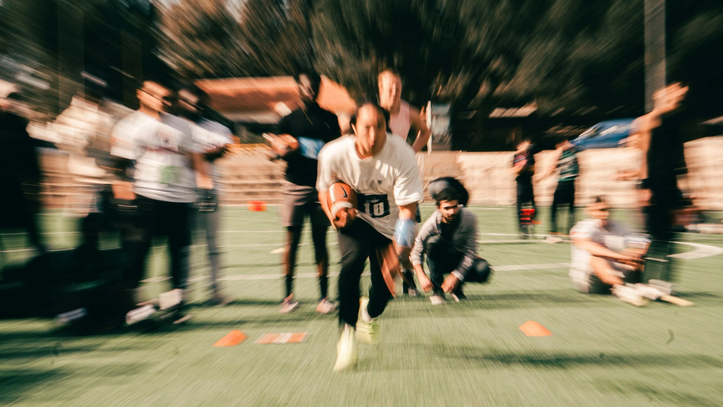 A person in a white shirt running with a football during a training drill on a football field, surrounded by several people, with a blurred background.