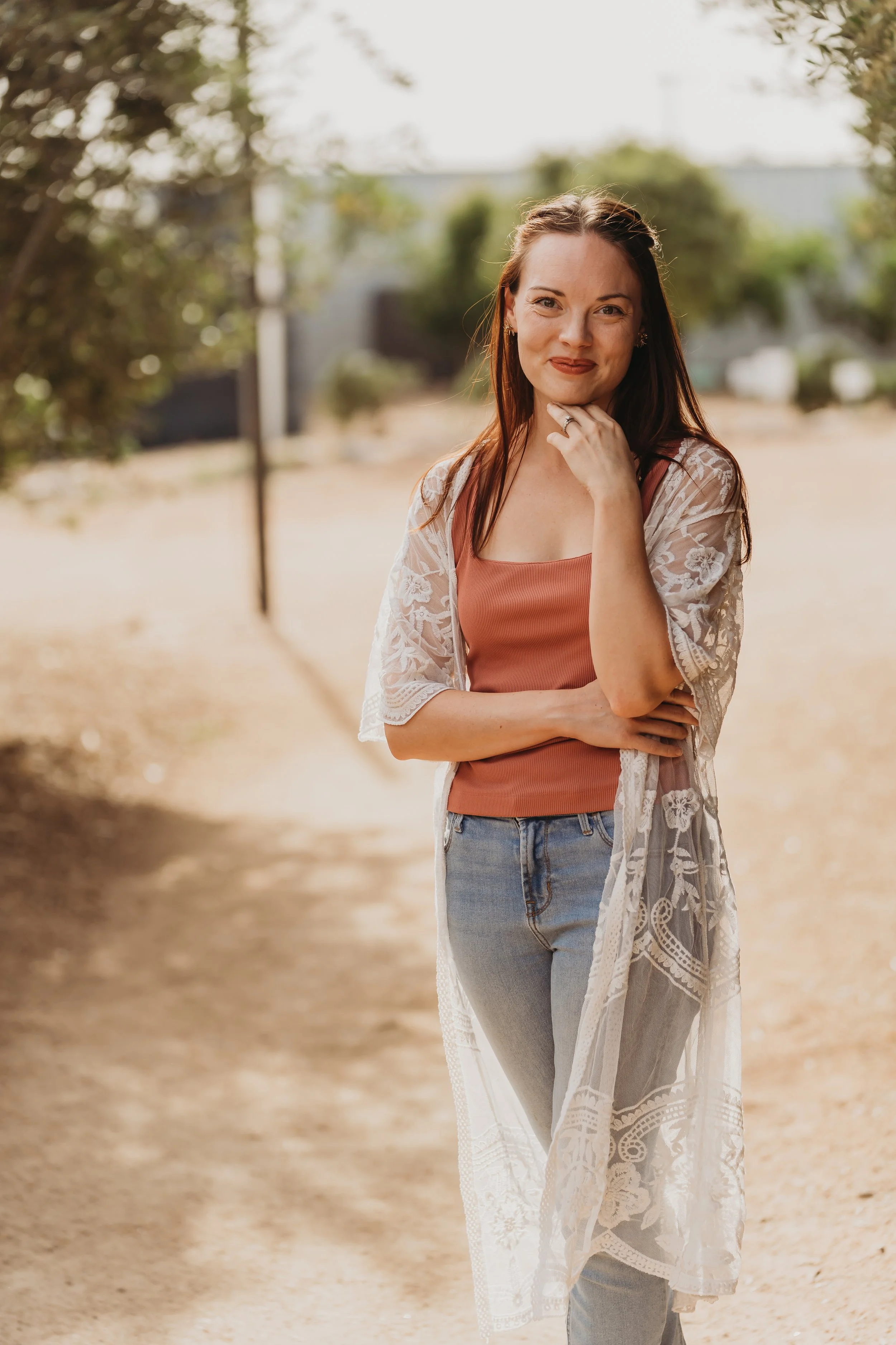 Postpartum doula Hannah standing on a dirt path outdoors, smiling, wearing a pink top, light blue jeans, and a white lace cover-up, with trees and a blurred background.