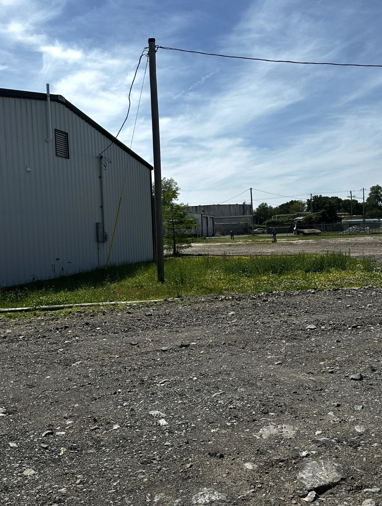 A gravel lot with patches of grass and a large white industrial building on the left. A utility pole with power lines, and a few distant structures and trees under a blue sky with scattered clouds.