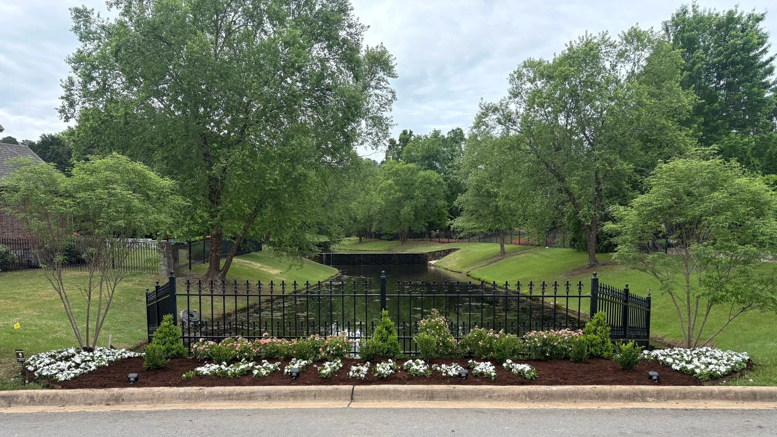 A landscaped area with a small water feature surrounded by flowers, shrubs, and trees, enclosed by a black metal fence, with a brick house partially visible on the left and a street in the foreground.