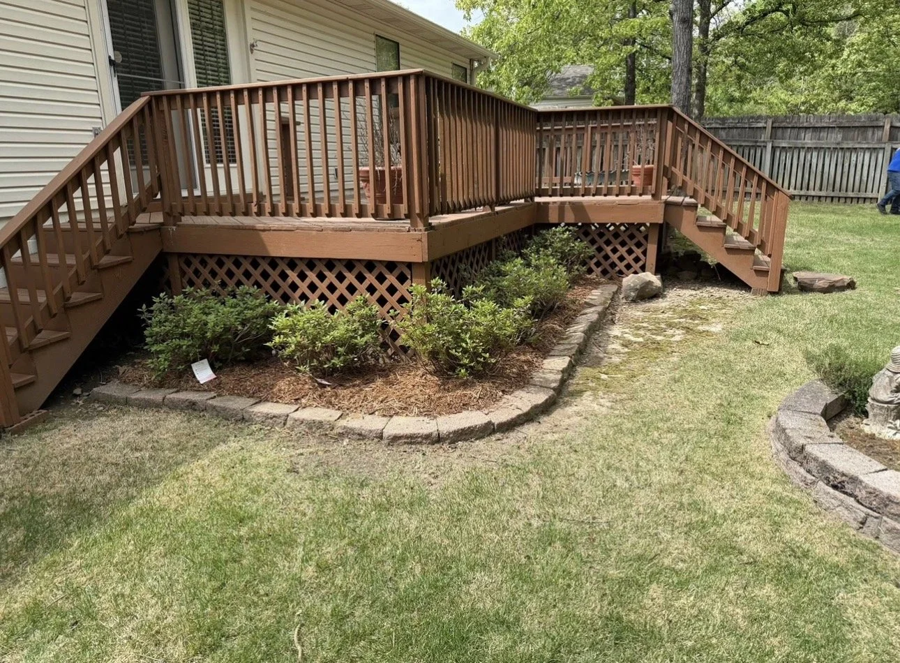 Backyard with a wooden deck with stairs and railing, green grass, small garden bed with plants, and a wooden fence in the background.
