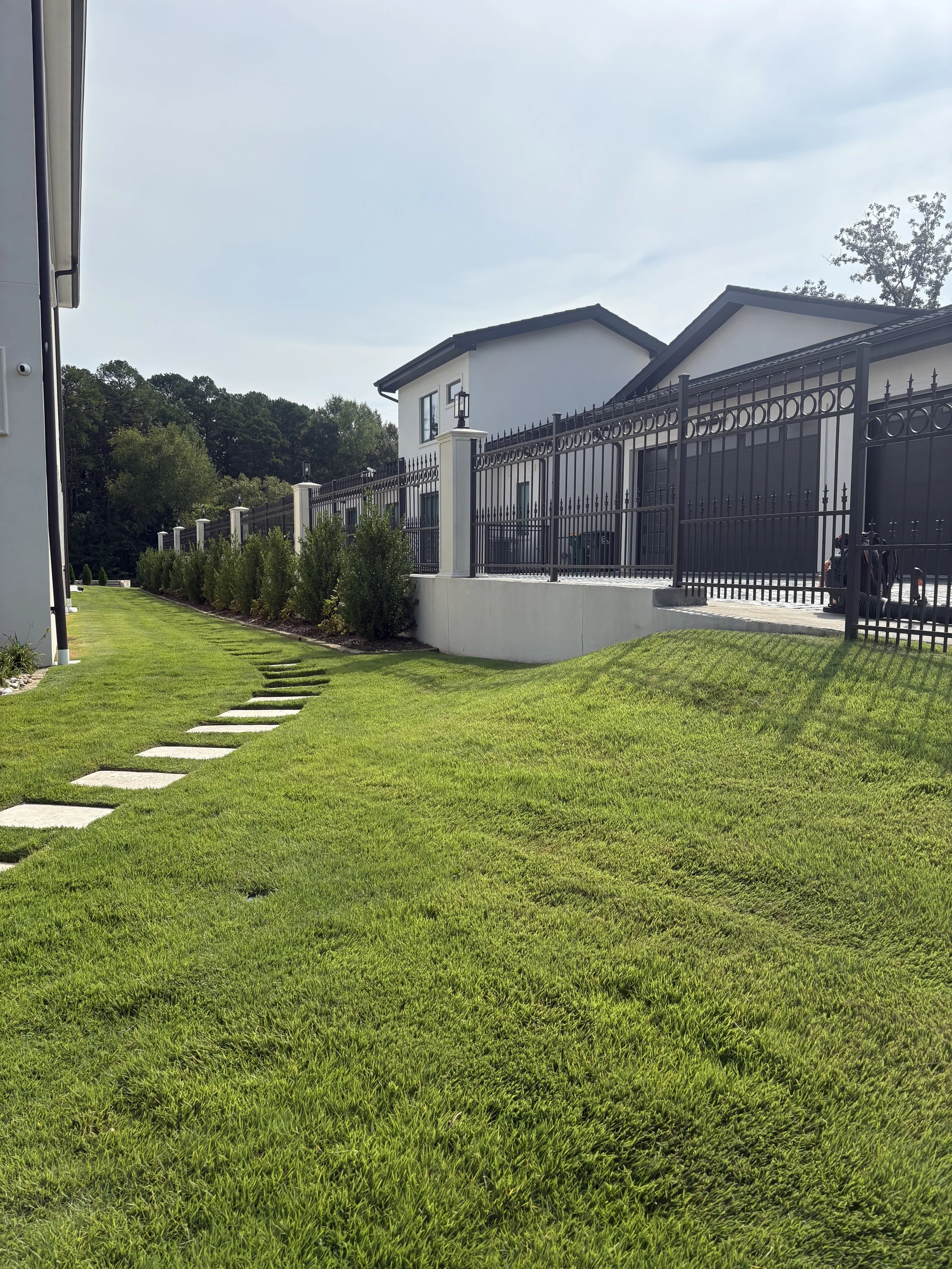 A well-maintained backyard with lush green grass, a stone pathway, and a white modern house with a black iron fence. There are trees in the background under a partly cloudy sky.