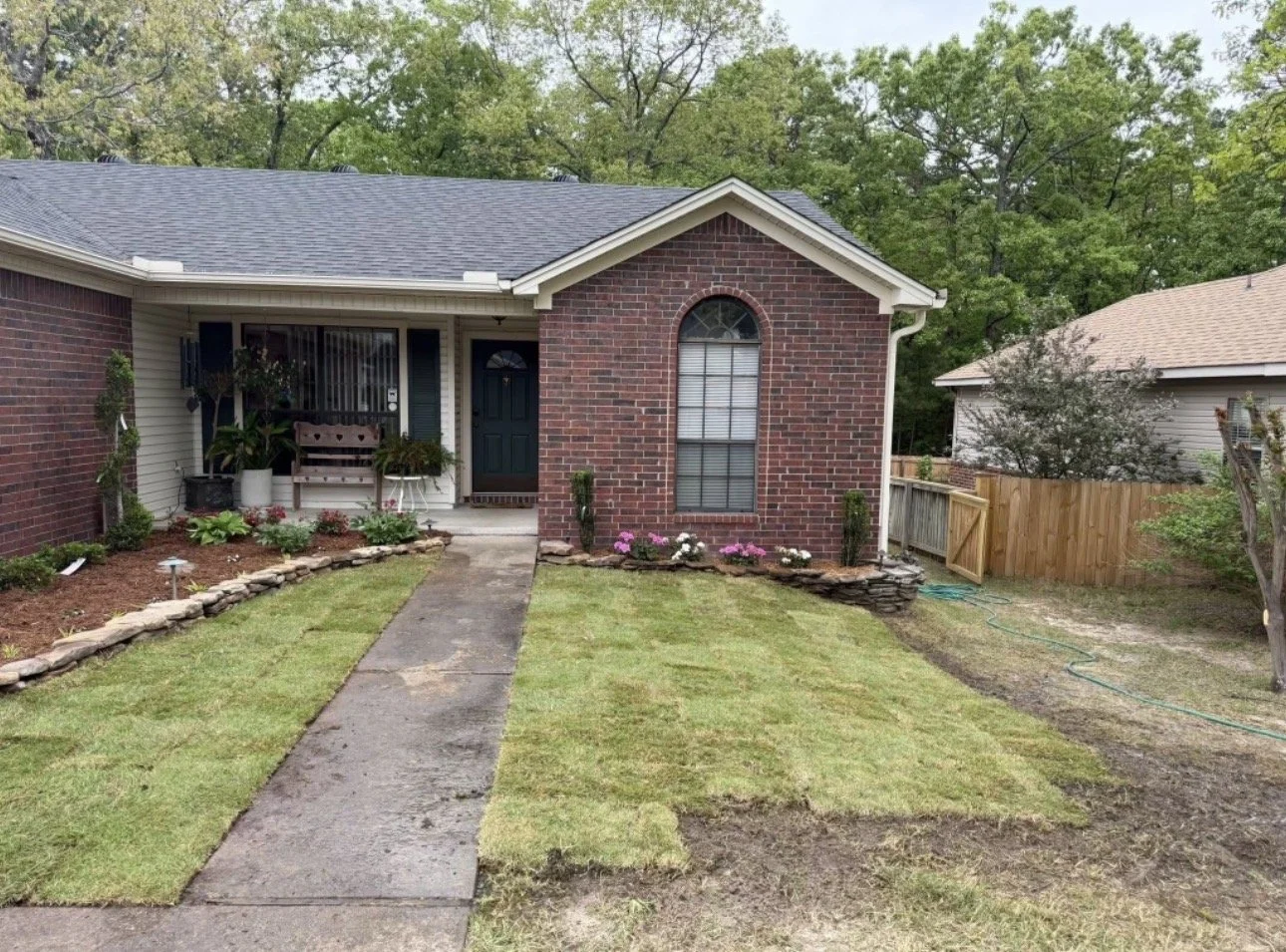 Front yard of a brick house with a walkway, flower bed, and a garden hose, surrounded by green trees in a suburban neighborhood.