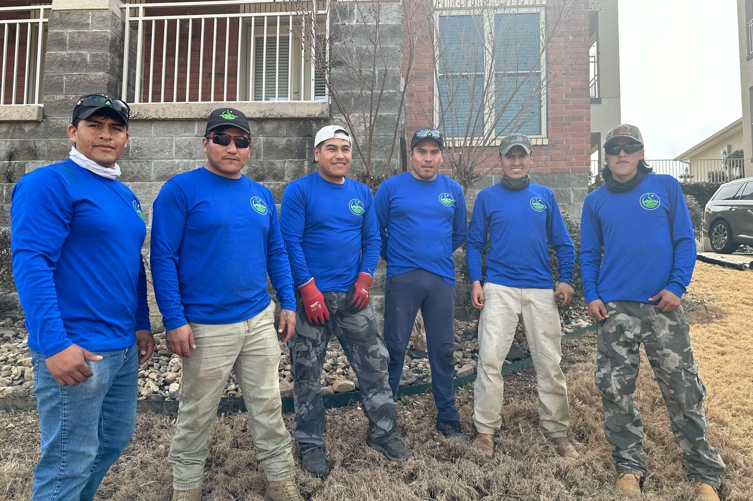 Seven men standing outdoors in front of a brick and stone building, wearing matching blue long-sleeve shirts with a logo, camouflage pants, and work gloves.