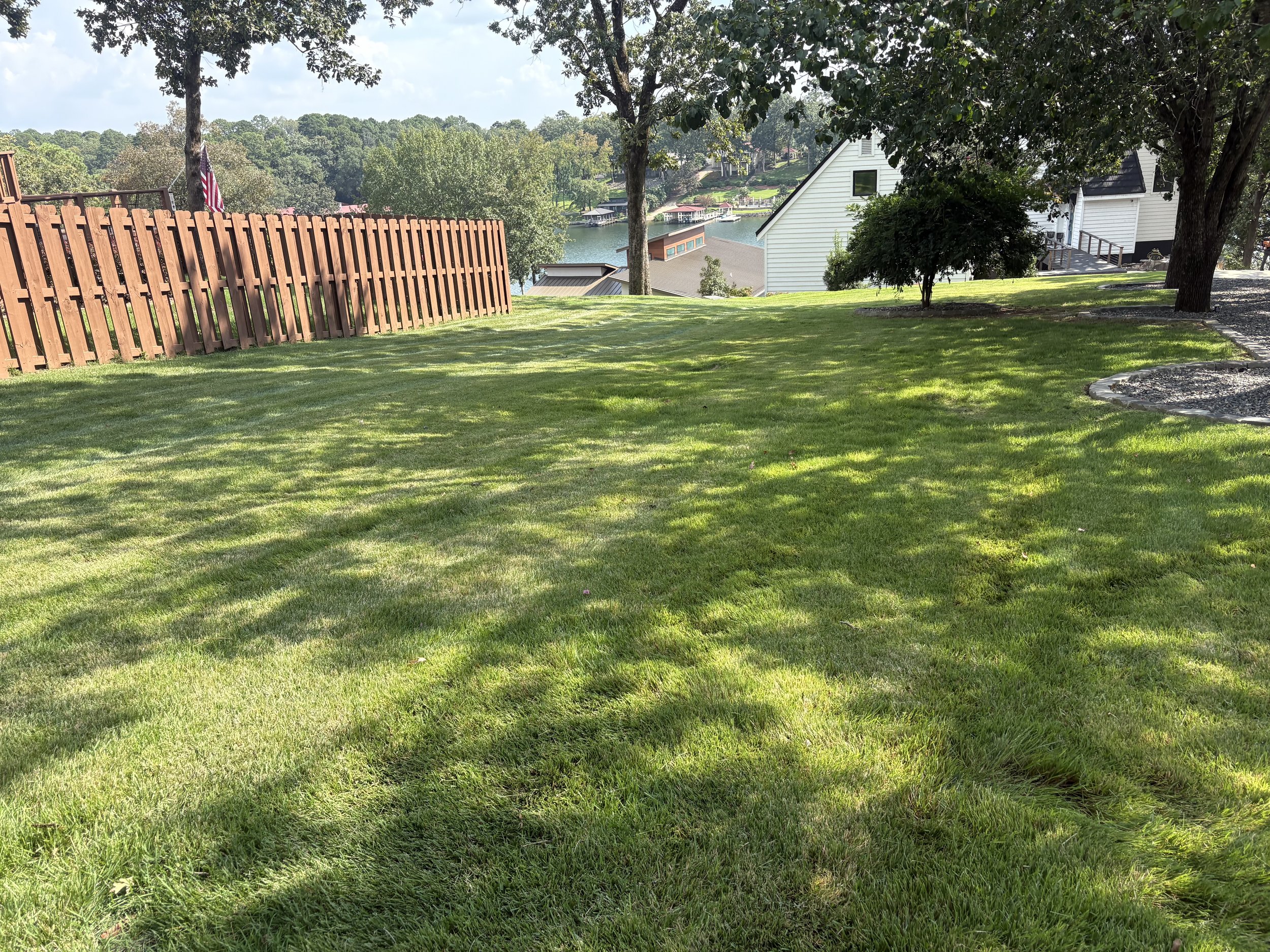 A lush green backyard with well-maintained grass, several large trees providing shade, a wooden fence on the left, and lakeside houses in the background near a body of water.