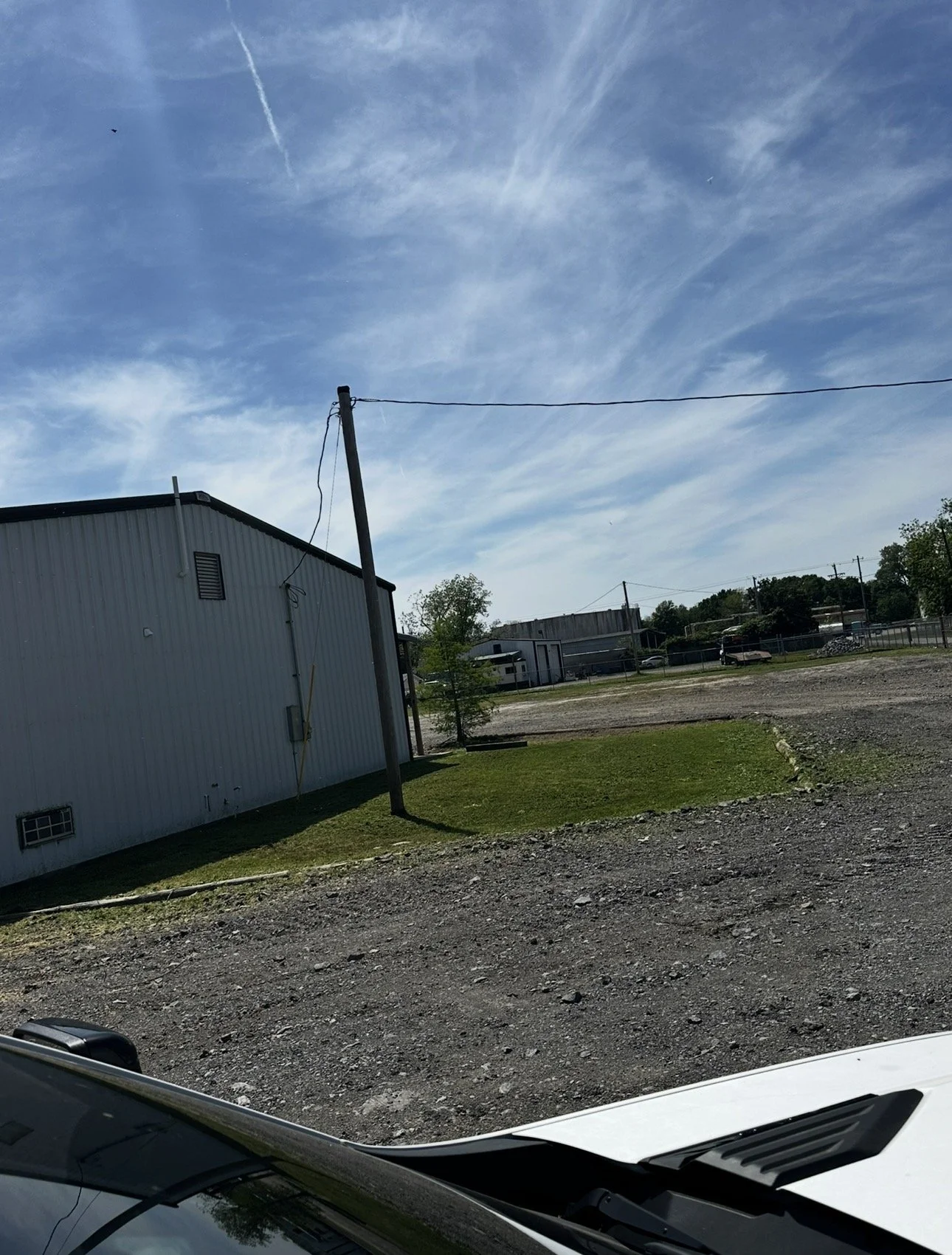View of a partially grassy and gravel lot with a white metal building, a leaning utility pole, and a bright blue sky with wispy clouds and airplane contrails.