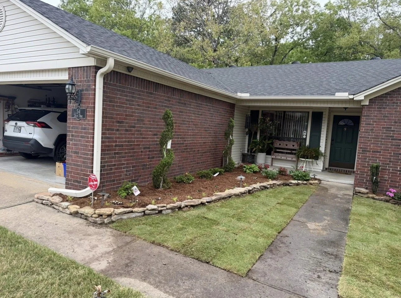 Front yard of a suburban house with a garden bed, walkway, and porch. The garden bed is bordered with rocks and has plants and shrubs, including two tall, twisted trees. The porch has a bench and potted plants, with the dark green front door visible.