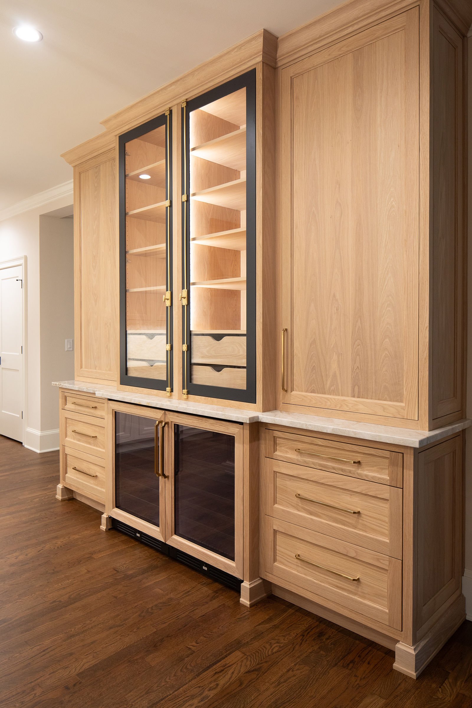 Built-in wooden cabinet with glass doors, drawers, and shelves, topped with a light-colored marble surface, located in a room with hardwood flooring.