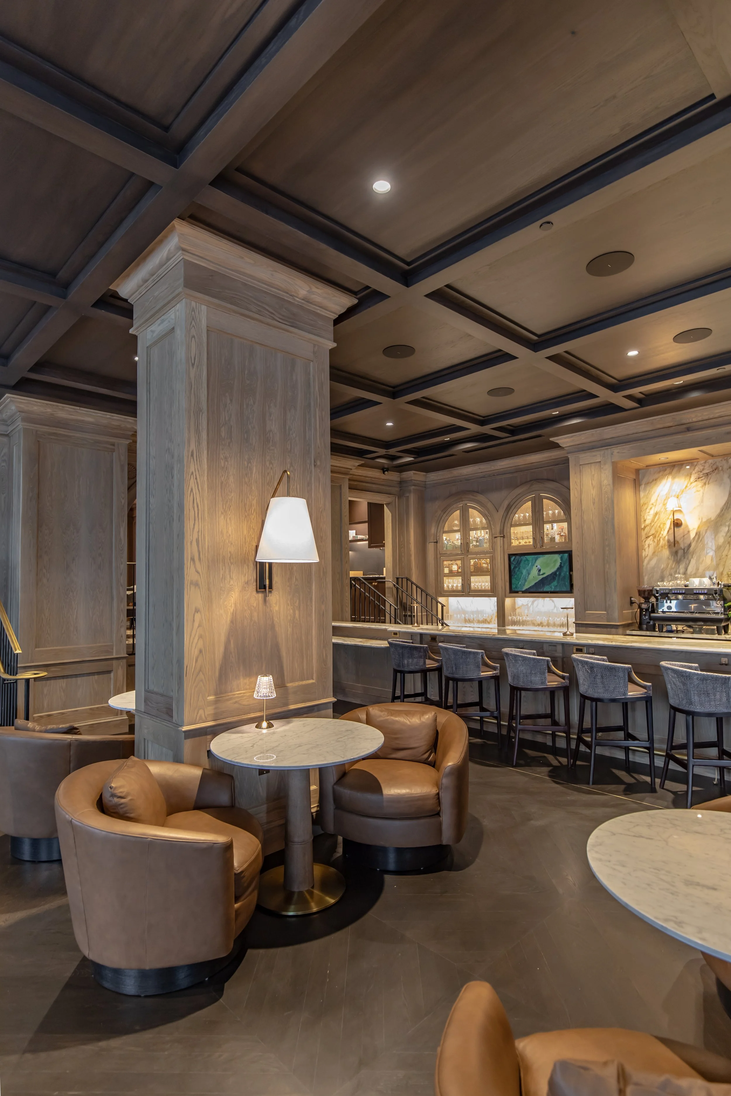 Interior of a modern restaurant or cafe with wooden paneling, leather chairs, marble tables, and a bar area with bar stools.