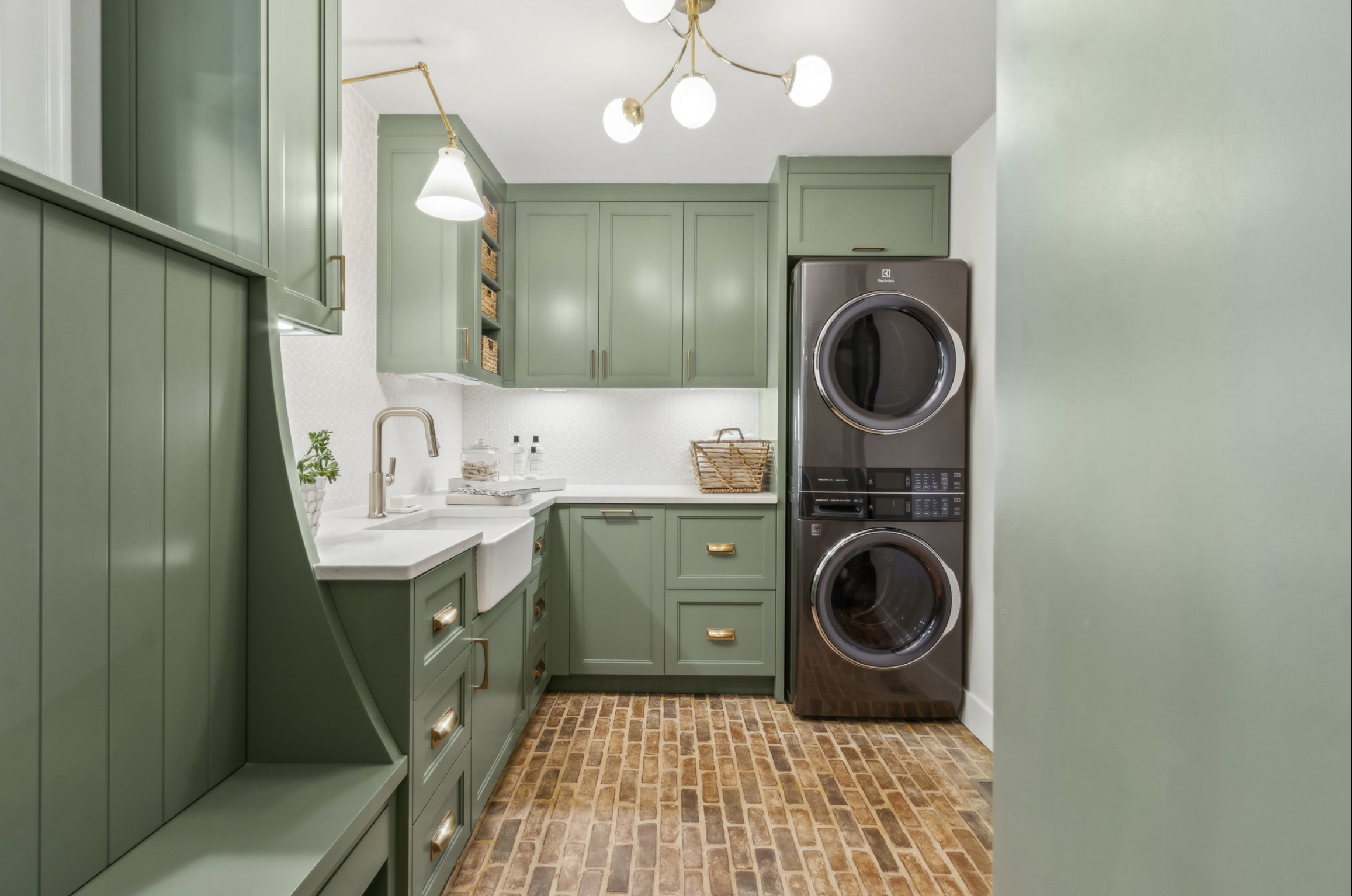 Laundry room with green cabinets, white countertop, a white sink with a bronze faucet, a basket, laundry supplies, and a stacked washer and dryer in stainless steel. The floor is made of brick-like tiles, and the room features a modern chandelier wit