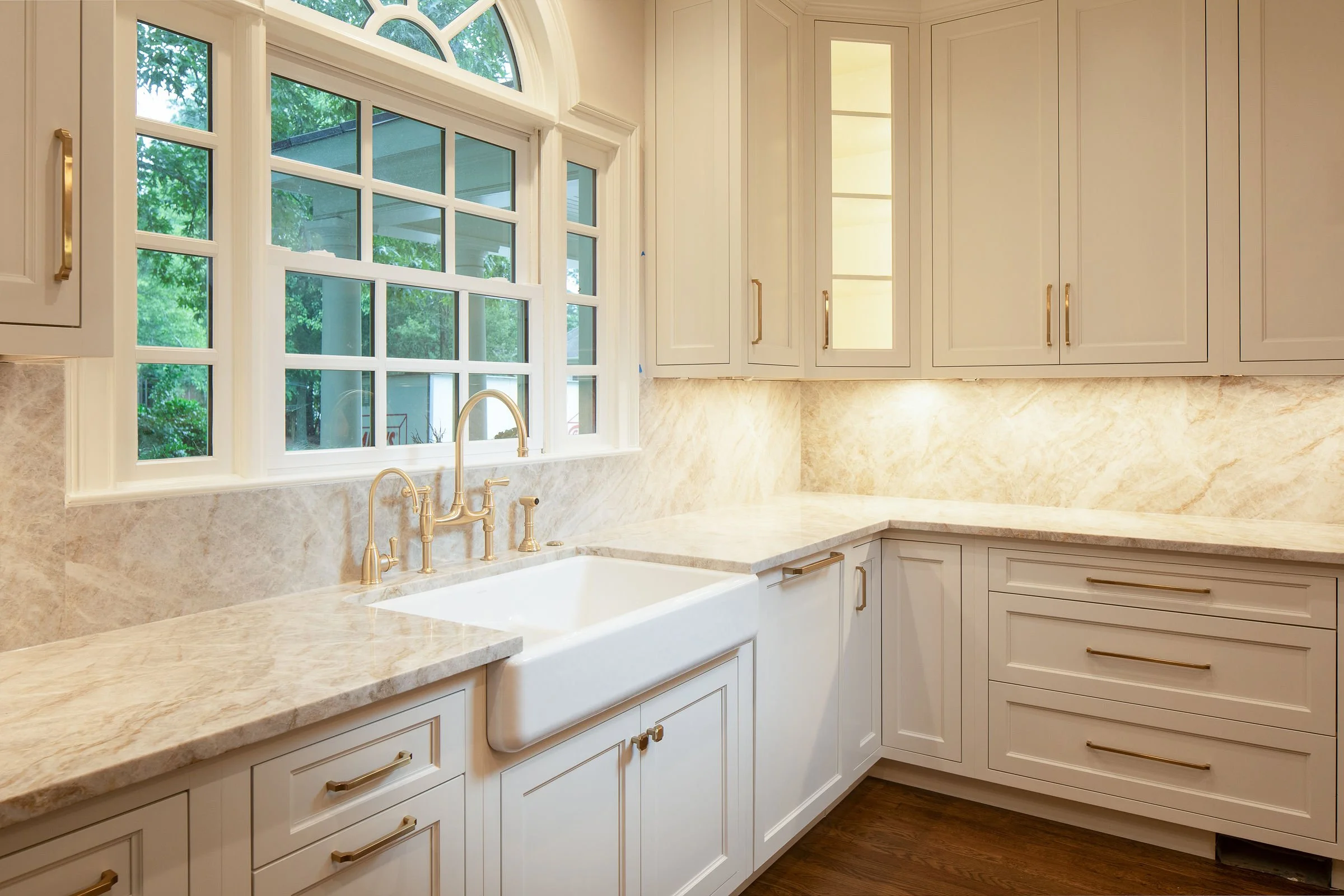 Kitchen with white cabinets, beige marble countertops, a large window above the sink, and brass fixtures.